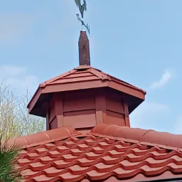 Czerwony dach z metalowej dachówki falistej, zwieńczony małą wieżyczką z wiatrowskazem w kształcie koguta, na tle błękitnego nieba.