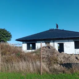 Czarny dach z dachówki ceramicznej na surowym domu z betonu komórkowego, pracownik na dachu montuje kominki wentylacyjne, w tle błękitne niebo i drzewo.