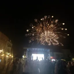 Nocny pokaz pirotechniczny nad sceną z występującymi artystami, tłum widzów obserwuje fajerwerki na tle oświetlonych budynków.