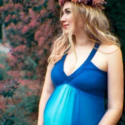 Kobieta w ciąży w niebieskiej sukience z kwiatowym wiankiem na głowie, spoglądająca w lewo na tle rozmytej zieleni i różowych kwiatów.