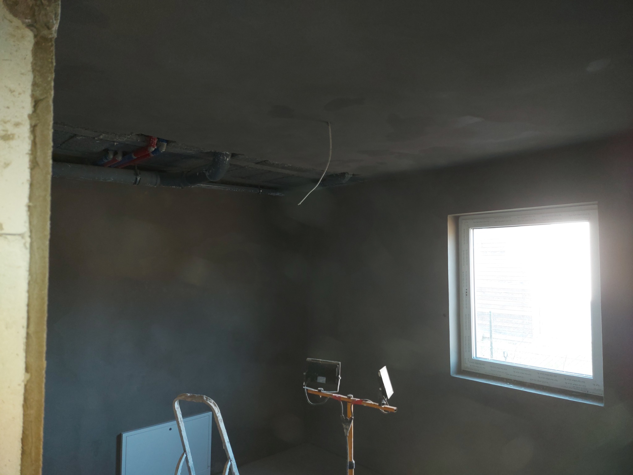 Pokój w trakcie remontu z świeżo nałożoną, szarą warstwą tynku na ścianach i suficie, widoczne rury instalacyjne pod sufitem, drabina i oświetlenie budowlane.