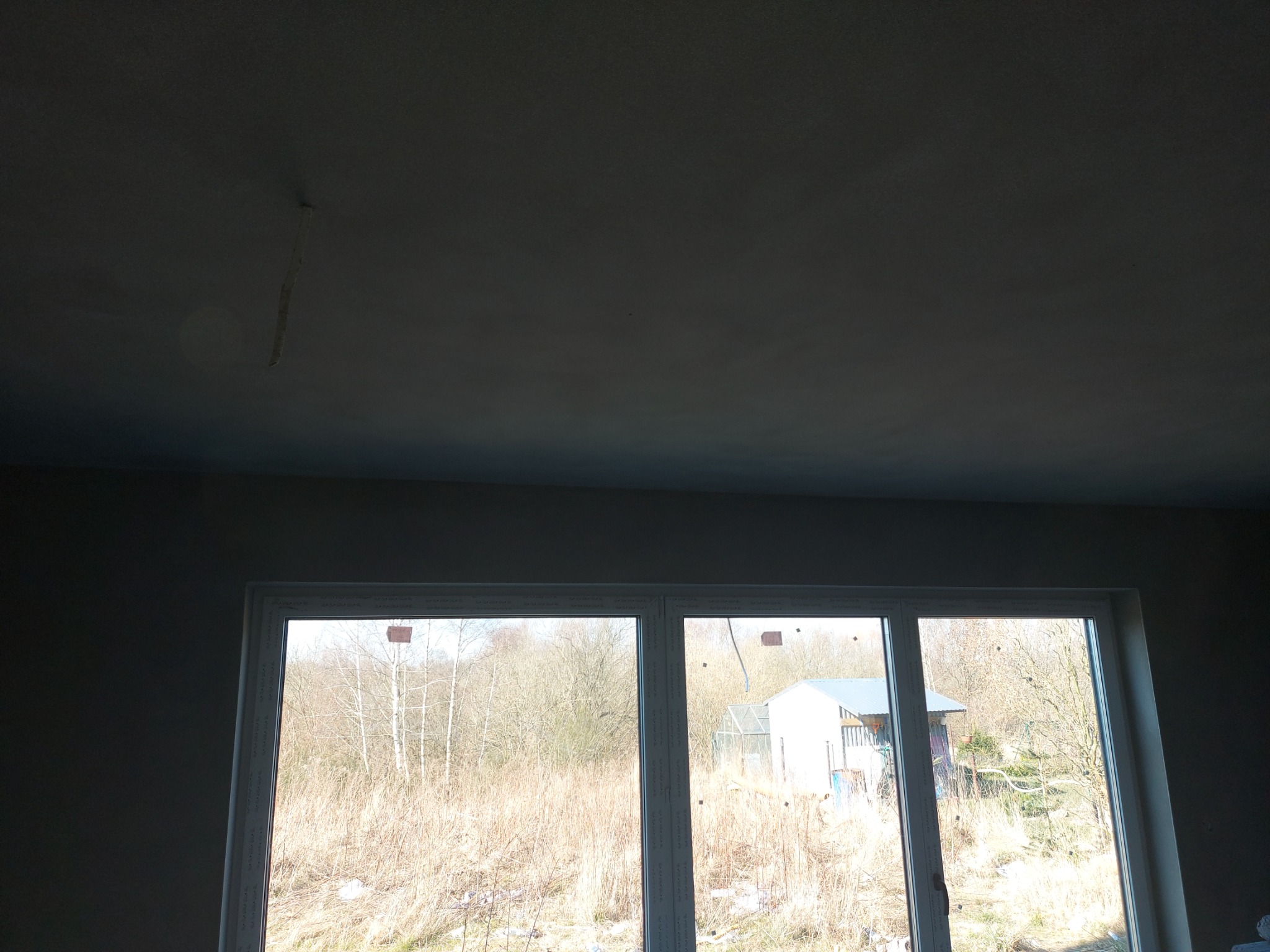 Świeżo otynkowany sufit z widocznymi niedociągnięciami i fragmentem zwisającego kabla, okno z widokiem na zarośnięte pole i budynek w tle.