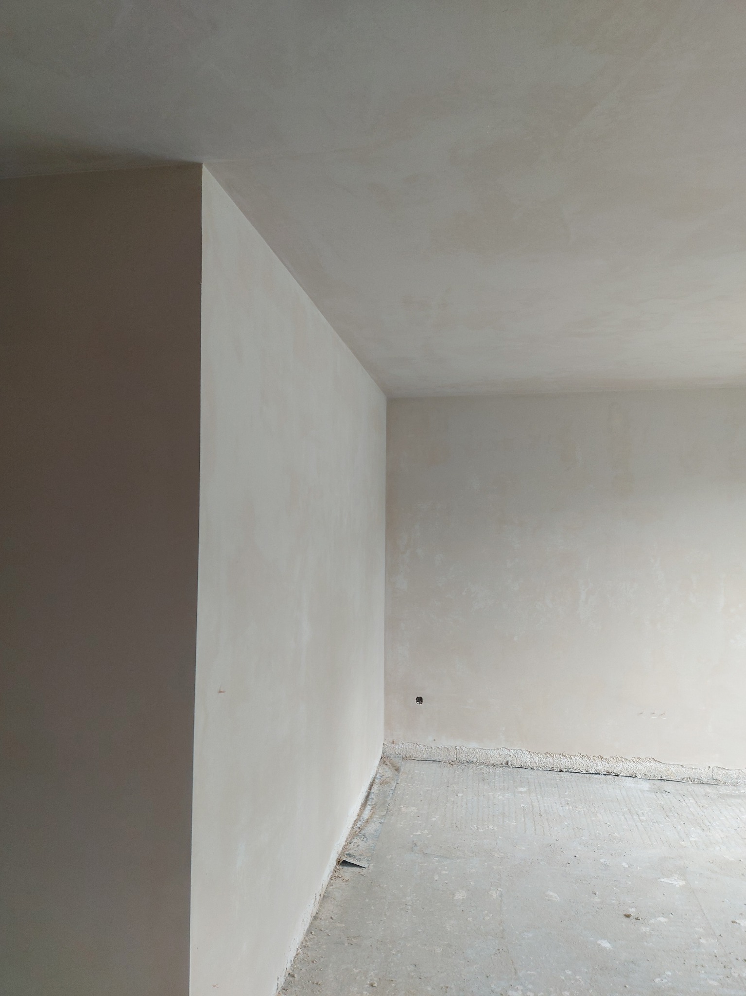 Wnętrze pokoju po nałożeniu gładzi gipsowej na ściany i sufit, widoczny surowy beton na podłodze i wystający narożnik ściany.