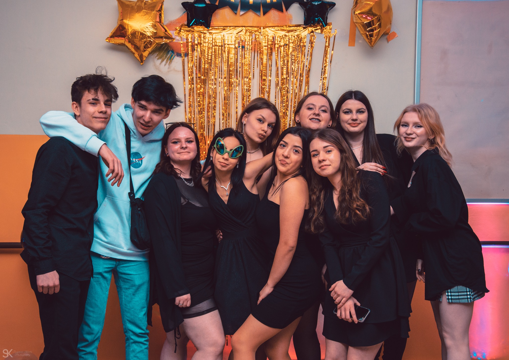 Grupa młodych ludzi pozuje do zdjęcia na tle złotej kurtyny i balonów w kształcie gwiazd podczas 18 urodzin Weroniki. Jedna osoba ma na sobie okulary w kształcie kosmitów.