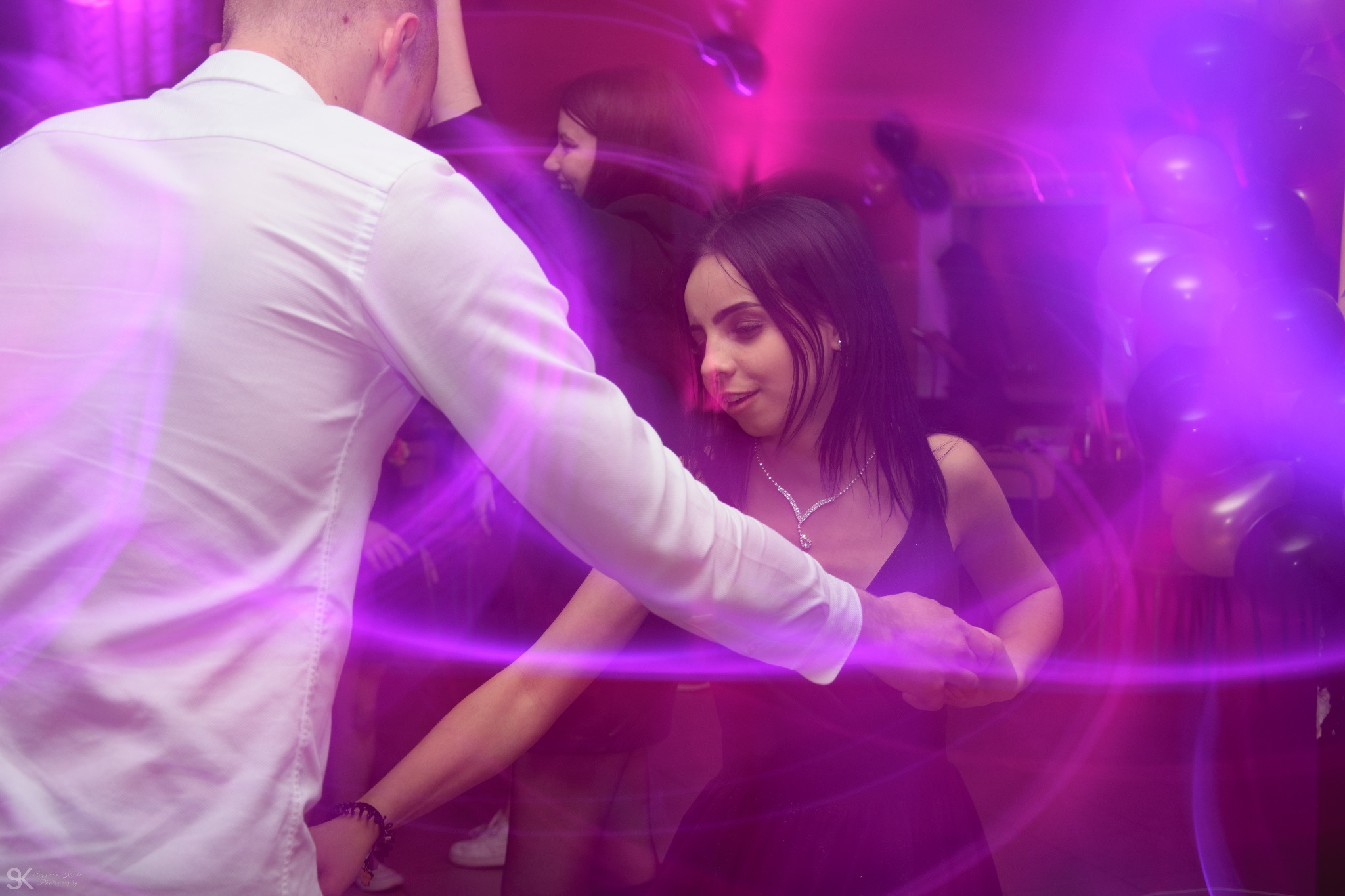 Młoda kobieta w czarnej sukience i z naszyjnikiem tańczy z mężczyzną w białej koszuli podczas przyjęcia urodzinowego, widoczne balony i efekty świetlne w kolorze fioletowym.