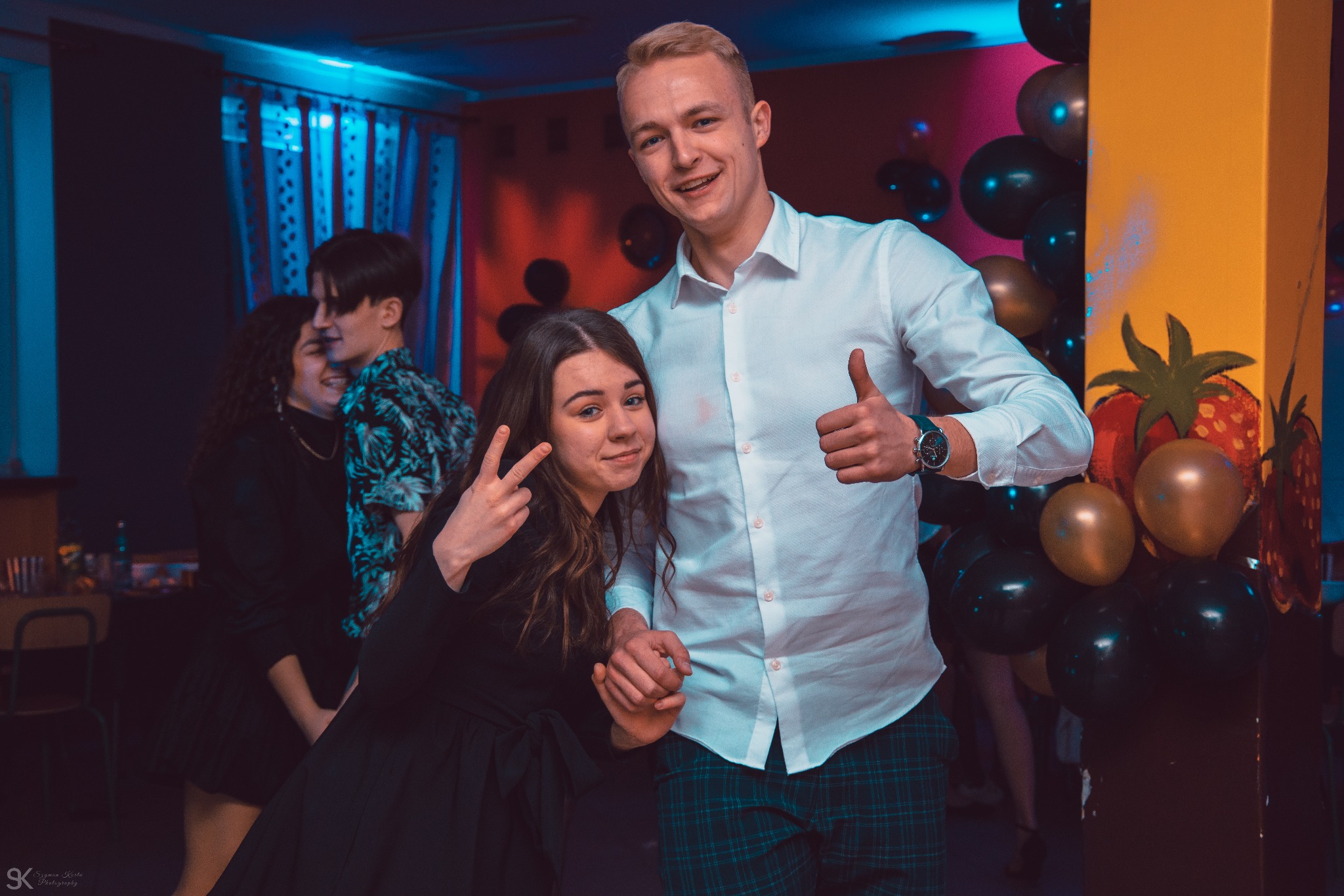 Uśmiechnięta dziewczyna w czarnej sukience pokazuje znak V, obok stoi chłopak w białej koszuli i zielonych spodniach w kratę, unosząc kciuk w górę, w tle dekoracje urodzinowe z balonów i kolorowy...