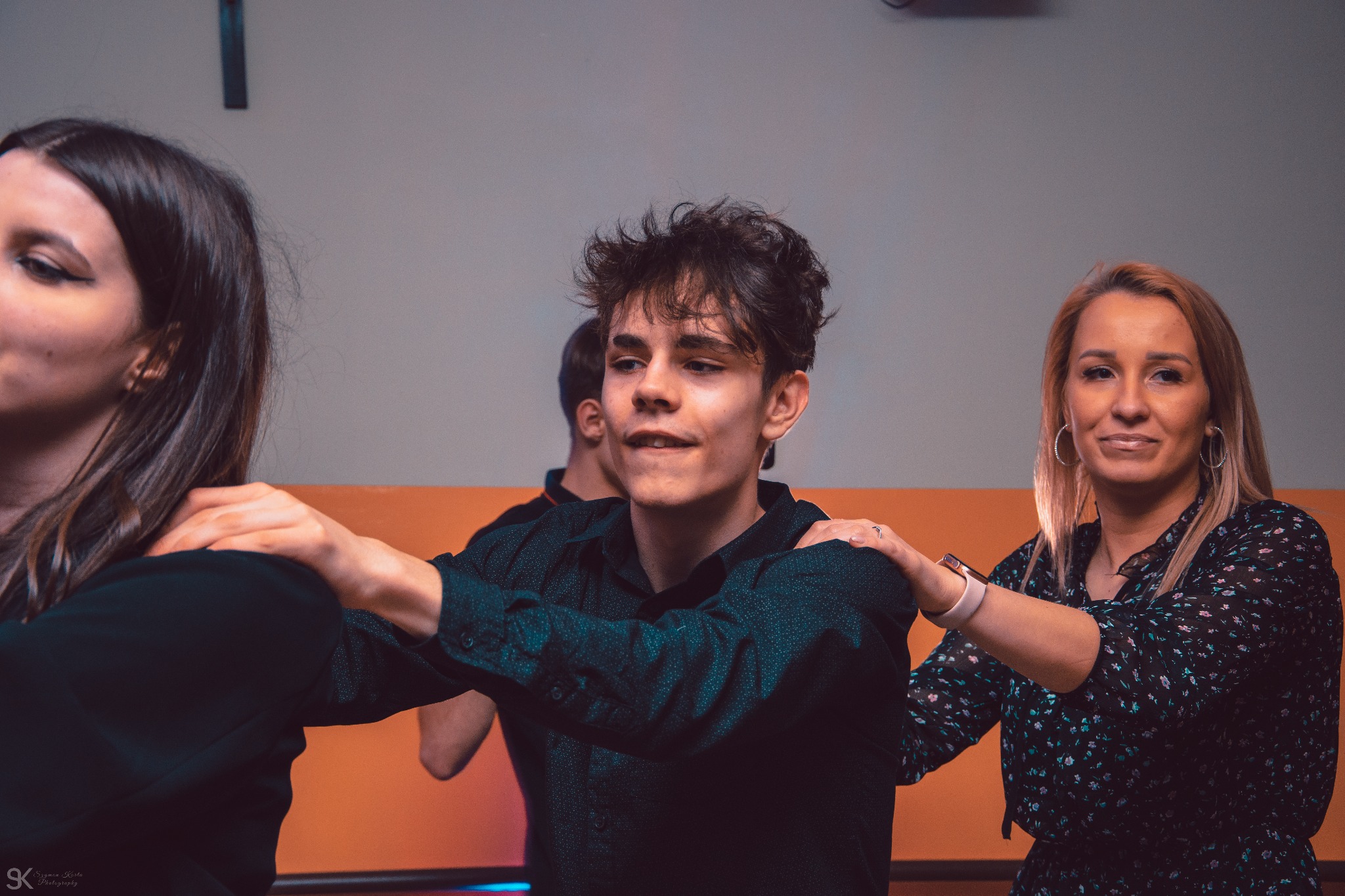 Młody mężczyzna w ciemnej koszuli, otoczony przez dwie kobiety, trzymające ręce na jego ramionach podczas zabawy na osiemnastych urodzinach Weroniki.