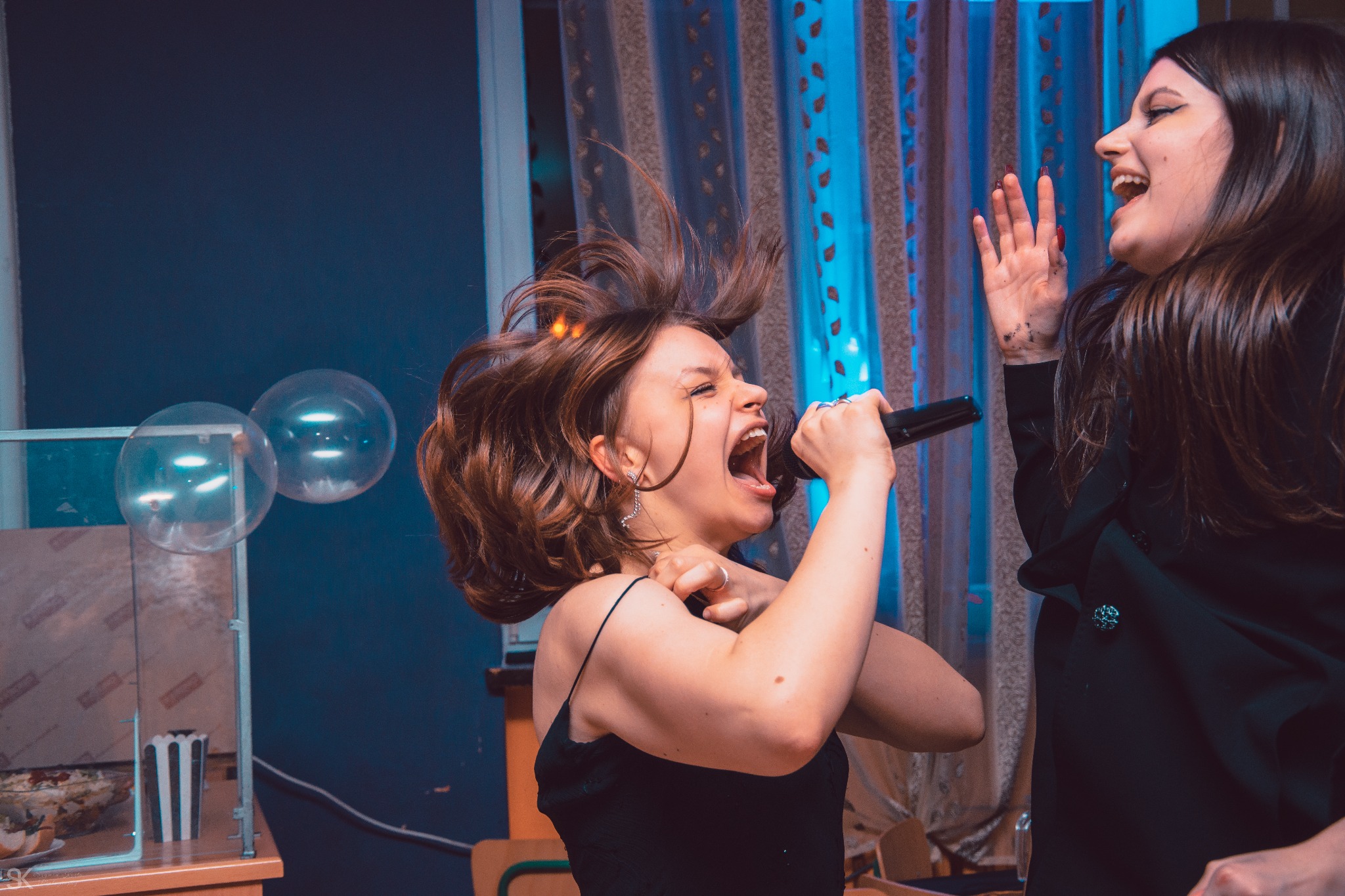 Rozentuzjazmowana młoda kobieta z mikrofonem śpiewa podczas imprezy urodzinowej; w tle balony i kobieta w czarnym żakiecie.