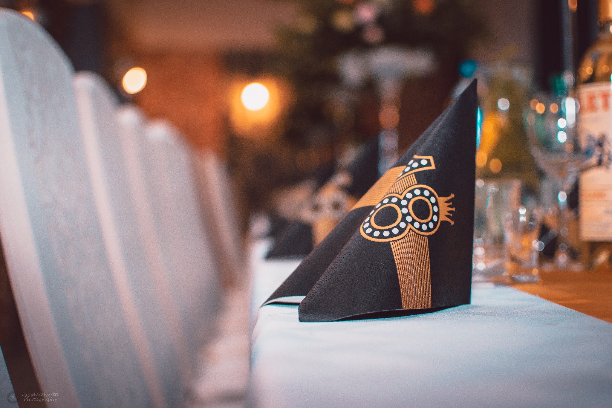 Dekoracyjny, czarny serwetnik z nadrukiem cyfry '18' w kolorze złotym, leżący na białym obrusie, w tle zastawiony stół z butelkami i kieliszkami, widok z perspektywy gościa siedzącego przy stole.