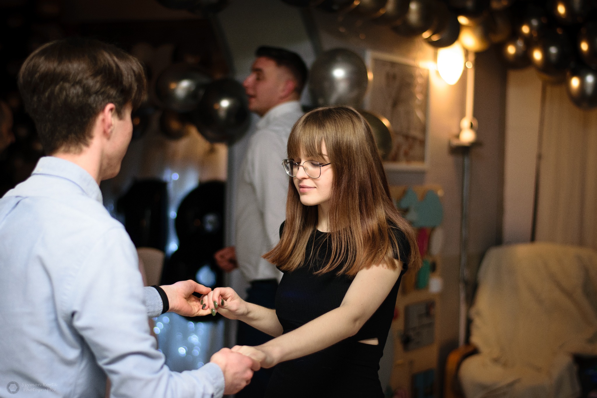 Młoda kobieta w czarnej sukience i okularach trzymająca dłoń mężczyzny podczas tańca na tle dekoracji z czarnych i srebrnych balonów na przyjęciu urodzinowym.