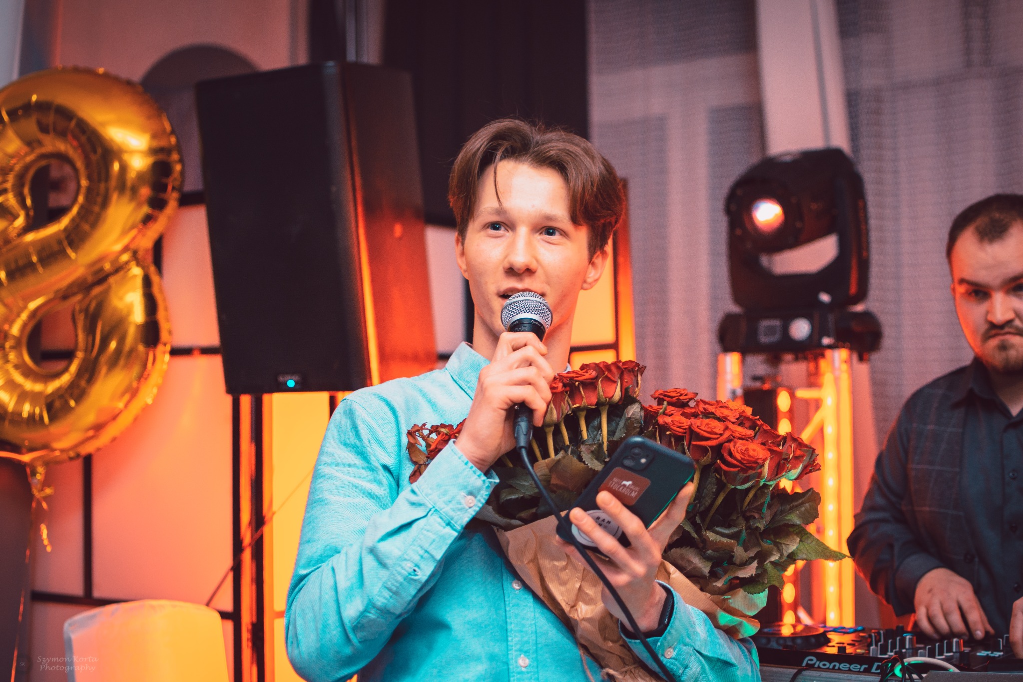 Młody mężczyzna w błękitnej koszuli trzyma mikrofon i bukiet czerwonych róż, w tle DJ przy konsolecie i dekoracje urodzinowe, w tym złoty balon w kształcie cyfry 8.