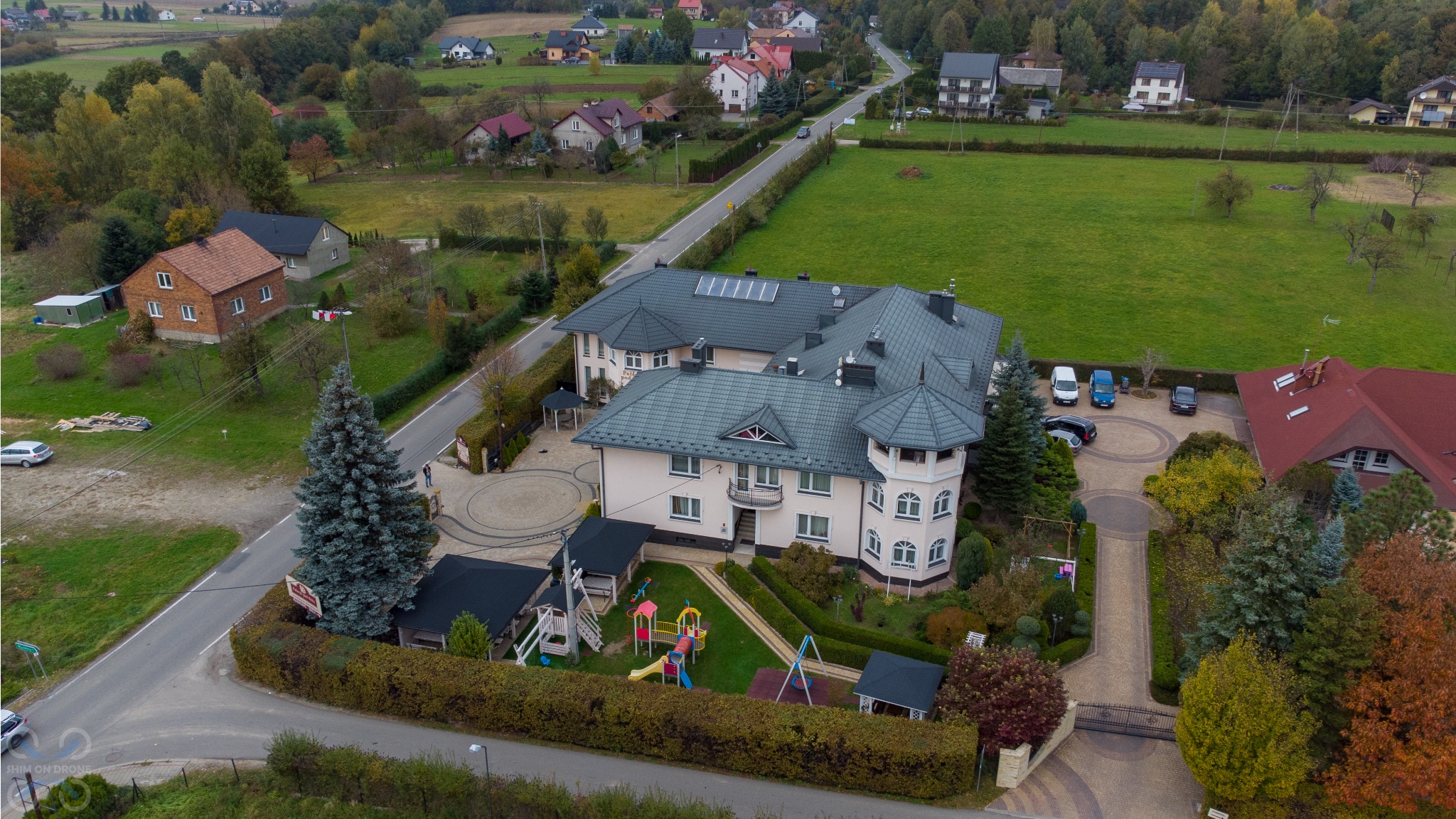 Widok z drona na duży, dwupiętrowy budynek z wieżyczką, otoczony zielenią, placem zabaw i parkingiem z kilkoma samochodami. Na dachu budynku widoczne panele słoneczne.