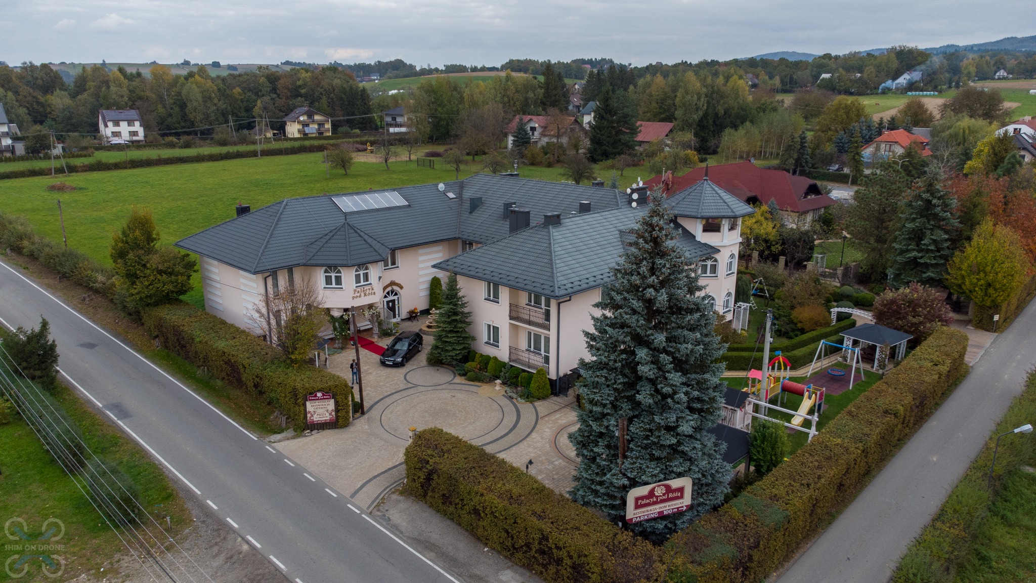 Widok z drona na budynek lokalu 'Pałacyk pod Różą' z szarym dachem, beżową elewacją, czerwonym dywanem przed wejściem, otoczony zielenią i placem zabaw.