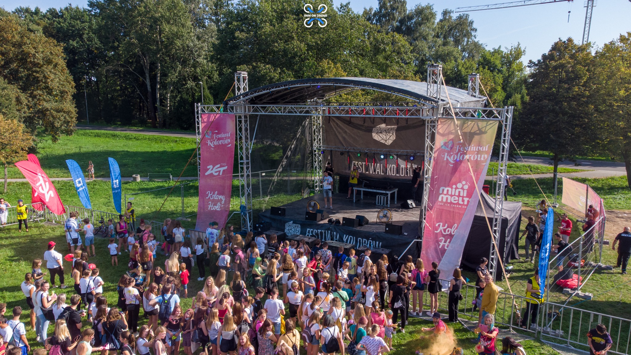 Widok z drona na scenę podczas Festiwalu Kolorów z tłumem uczestników, kolorowe flagi i banery reklamowe, scena z nagłośnieniem i oświetleniem, widok na park i drzewa w tle.