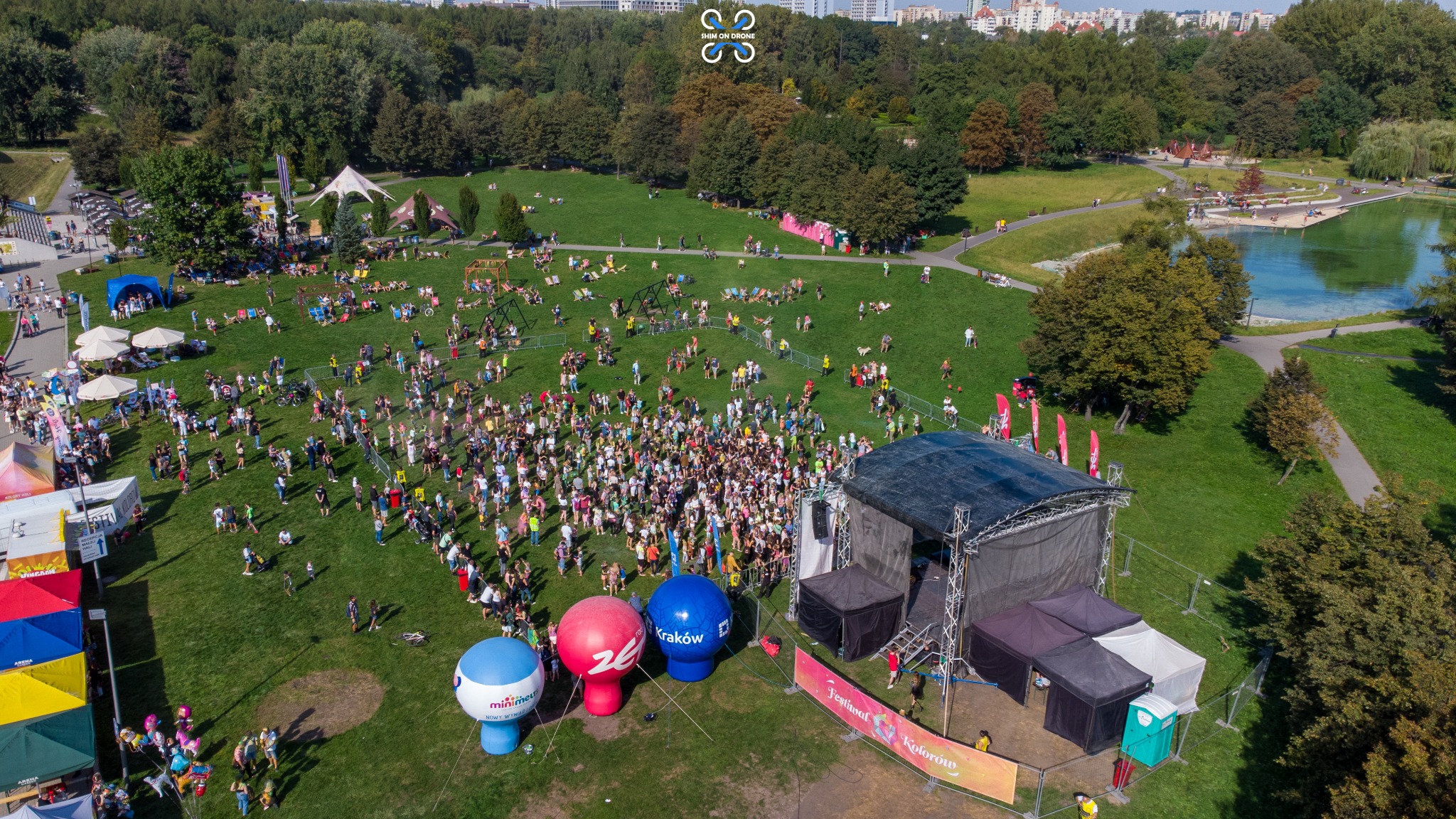 Widok z drona na Festiwal Kolorów w Krakowie: tłum ludzi na zielonej łące, scena, kolorowe balony reklamowe, park z jeziorem w tle.