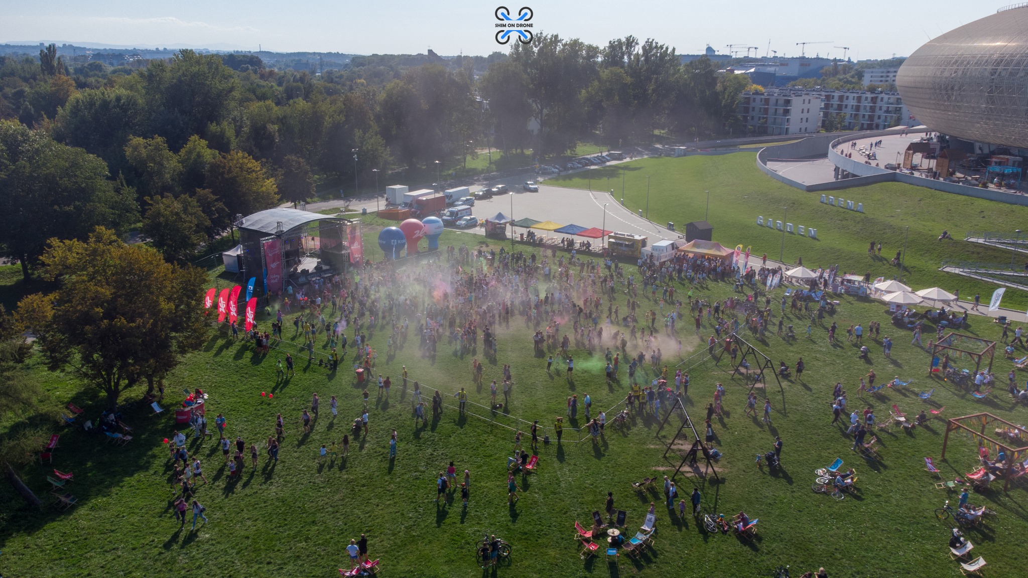 Widok z drona na Festiwal Kolorów na zielonej łące, z widoczną sceną, dmuchanymi balonami i licznymi uczestnikami rzucającymi kolorowymi proszkami, w tle drzewa i budynki.