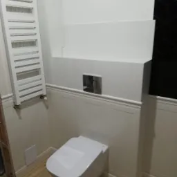 Nowoczesna toaleta wisząca w jasnej łazience z grzejnikiem drabinkowym i czarnymi płytkami w geometryczny wzór w rogu.