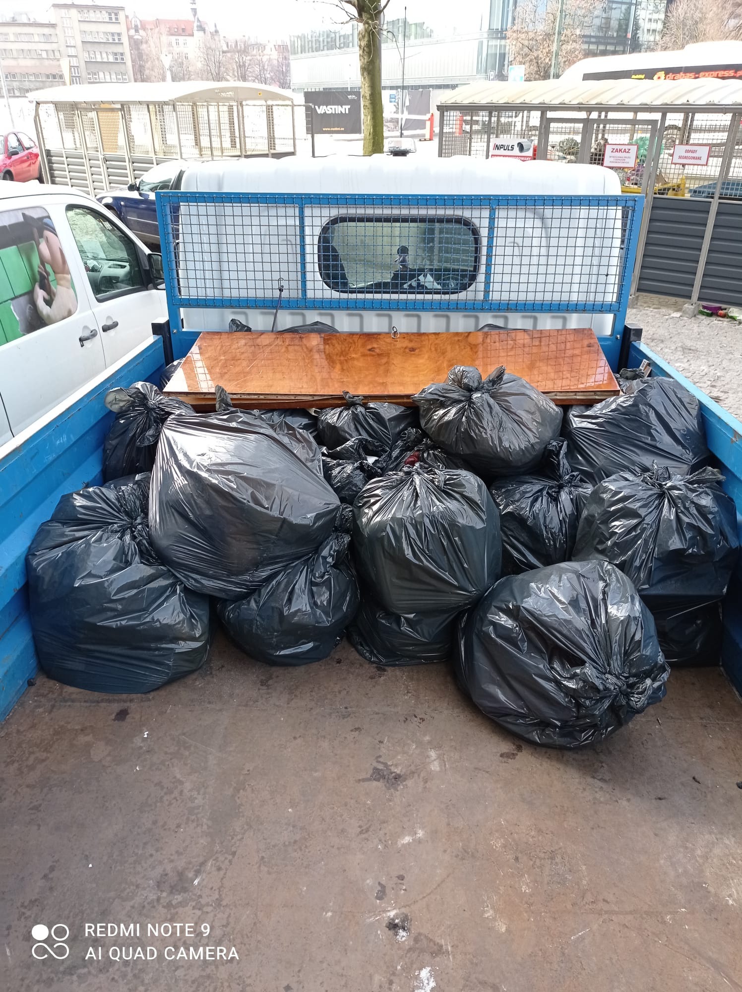 Pakaż ładunkowa pickupa wypełniona czarnymi workami na śmieci i drewnianymi panelami, widok z góry, w tle budynki miejskie.