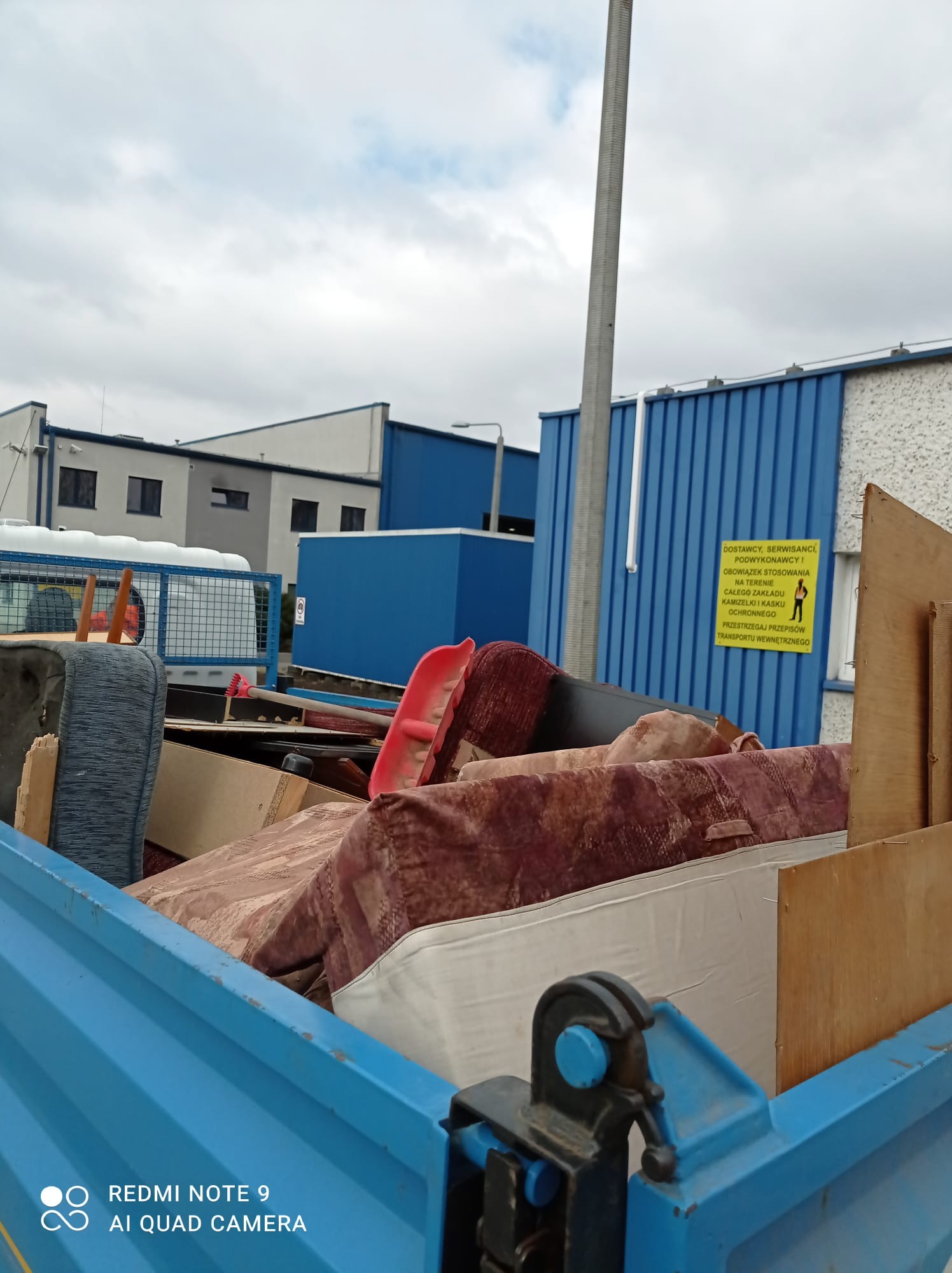 Załadunek starej sofy, materaca i mebli do niebieskiej przyczepy w przemysłowej scenerii z budynkami magazynowymi w tle.