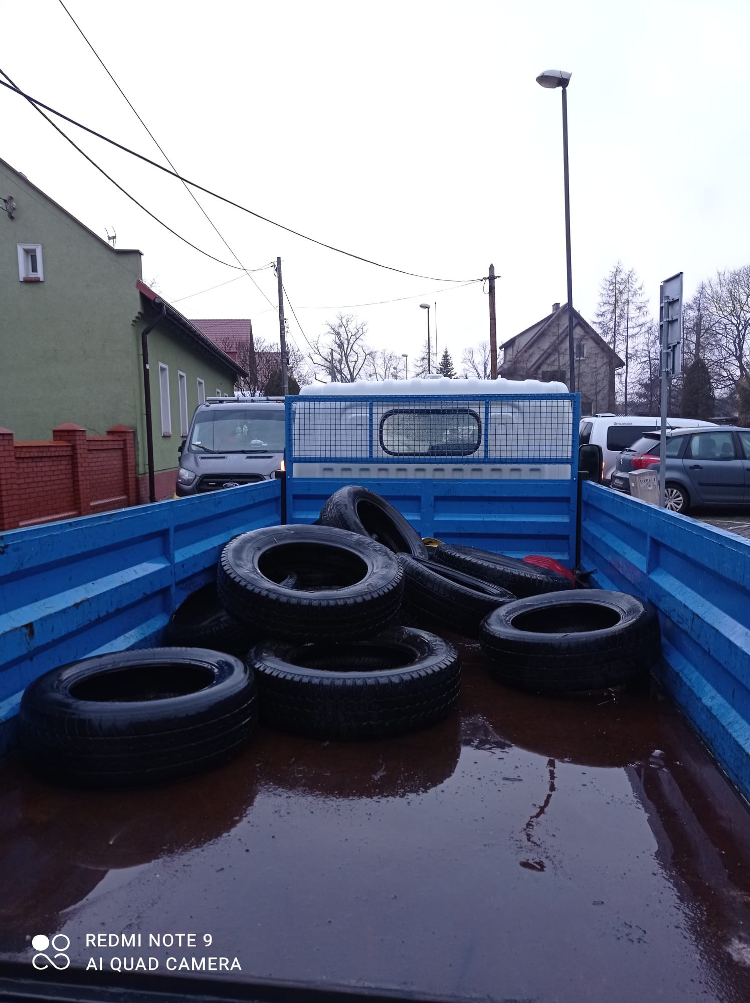Opony w skrzyni ładunkowej niebieskiego samochodu dostawczego, widok z góry na mokrą podłogę skrzyni, w tle budynki i zaparkowane samochody.