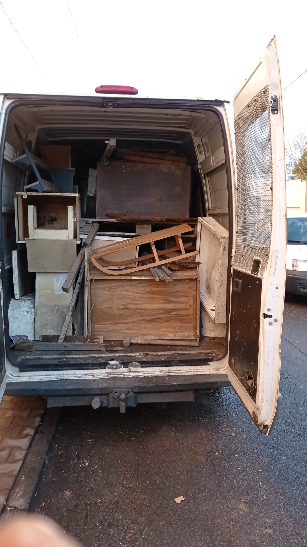 Wnętrze białego busa załadowane różnorodnymi przedmiotami, w tym drewnianymi skrzynkami, ramami i elementami konstrukcyjnymi, widoczne przez otwarte tylne drzwi z siatką ochronną.