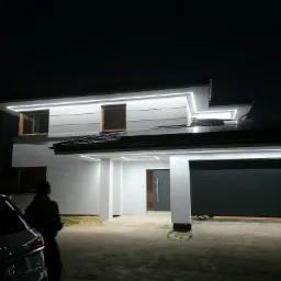 Nowoczesny dom jednorodzinny nocą z podświetloną elewacją LED, widoczne okna z żaluzjami, brama garażowa i fragment samochodu z sylwetką osoby.