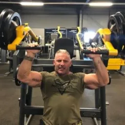 Mężczyzna z siwiejącymi włosami, wykonujący ćwiczenie na maszynie do wyciskania siedząc, w tle widoczna siłownia.