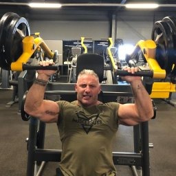 Mężczyzna z siwiejącymi włosami, wykonujący ćwiczenie na maszynie do wyciskania siedząc, w tle widoczna siłownia.