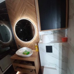 PERFEKT REMONTY I WYKOŃCZENIA - Nowoczesna łazienka z drewnianymi panelami ułożonymi w jodełkę, okrągłym lustrem z podświetleniem LED, wiszącą szafką z czarnymi frontami oraz umywalką w kształcie łzy i toaletą z marmurowym...