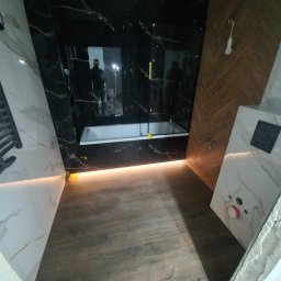 PERFEKT REMONTY I WYKOŃCZENIA - Nowoczesna łazienka w trakcie wykańczania: czarne płytki imitujące marmur, drewnopodobne panele na podłodze, biała wanna i podświetlenie LED, widoczne odbicie osoby robiącej zdjęcie w lustrze.
