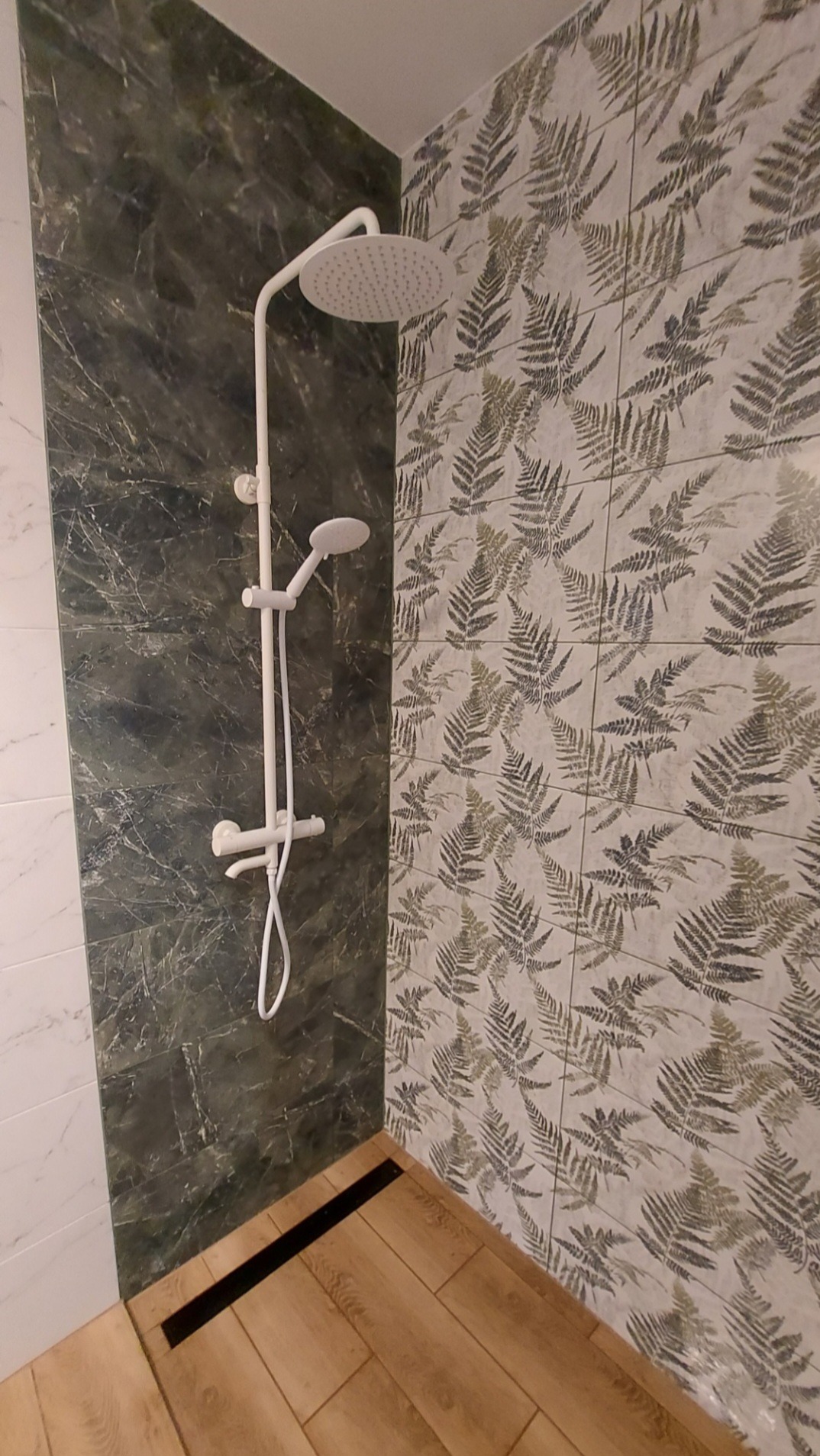 Nowoczesna kabina prysznicowa z deszczownicą i panelami ściennymi: marmurkowy szary i wzór liści paproci, drewnopodobna podłoga z czarnym odpływem liniowym.