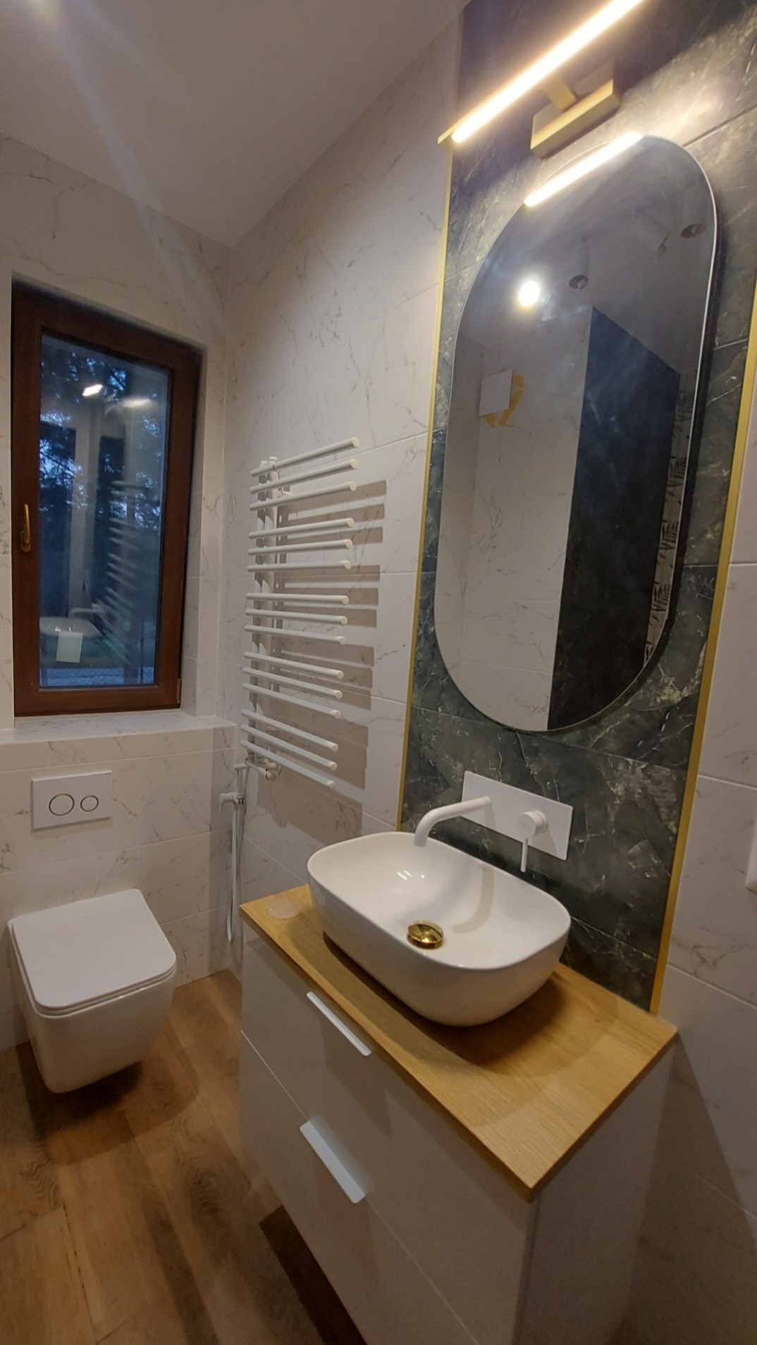 Nowoczesna łazienka z umywalką nablatową, podwieszaną toaletą i grzejnikiem drabinkowym na tle marmurowych płytek.