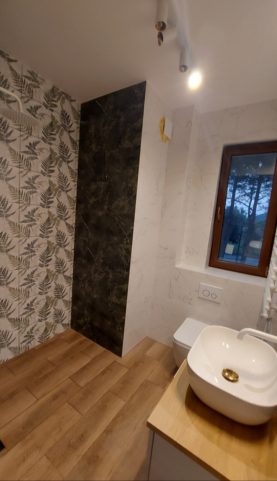 Nowoczesna łazienka z drewnopodobną podłogą, białą umywalką nablatową z złotym korkiem, płytkami ściennymi imitującymi marmur i liście paproci oraz oknem z widokiem na zieleń.
