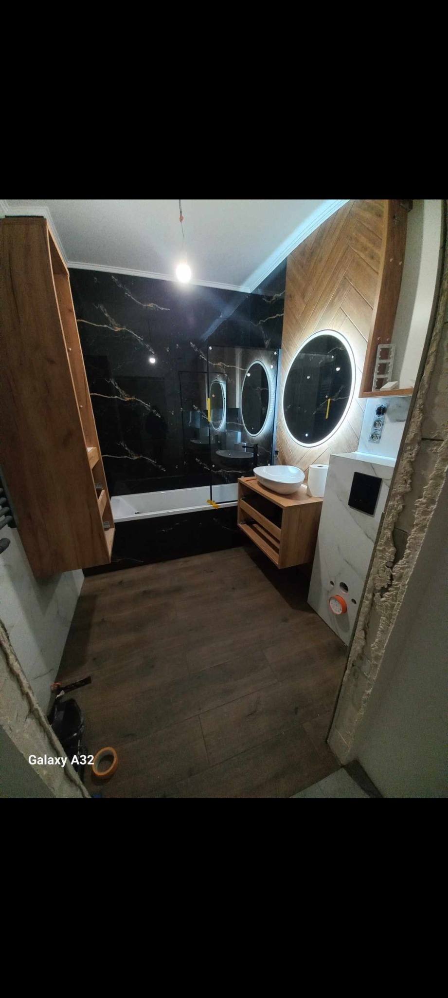 Nowoczesna łazienka w trakcie wykańczania, z czarnymi płytkami imitującymi marmur, drewnianą zabudową, okrągłymi lustrami z podświetleniem LED i podłogą imitującą drewno, widoczne niedokończone...