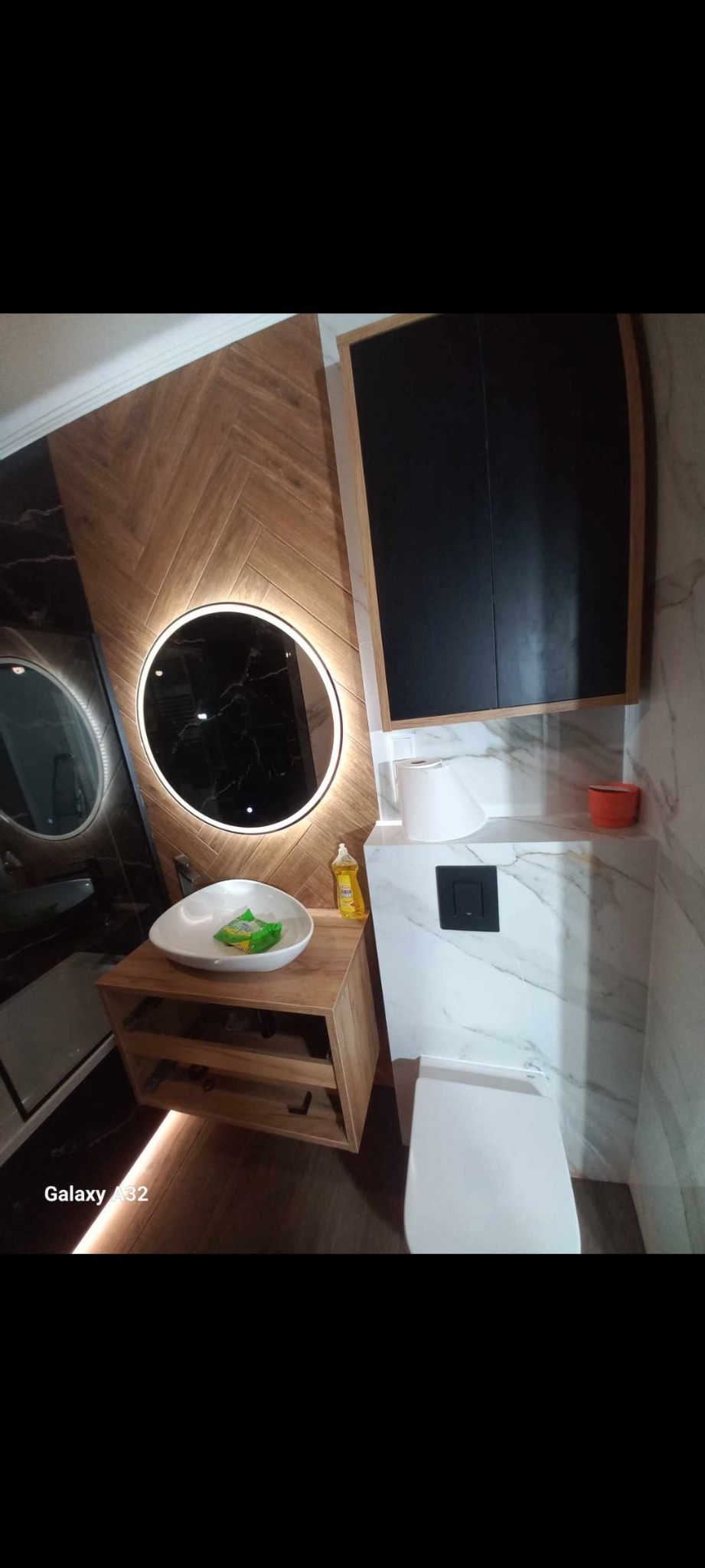 Nowoczesna łazienka z drewnianymi panelami ułożonymi w jodełkę, okrągłym lustrem z podświetleniem LED, wiszącą szafką z czarnymi frontami oraz umywalką w kształcie łzy i toaletą z marmurowym...