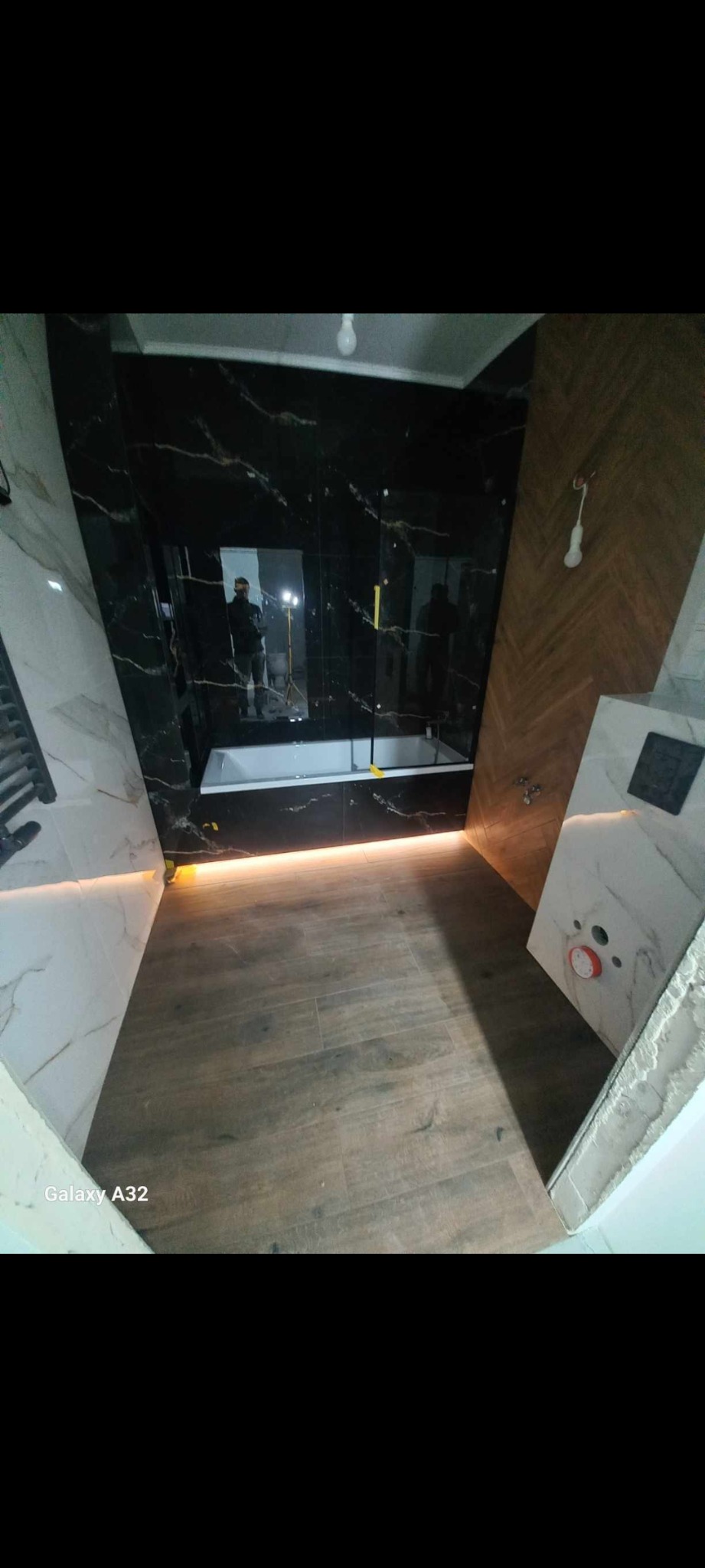 Nowoczesna łazienka w trakcie wykańczania: czarne płytki imitujące marmur, drewnopodobne panele na podłodze, biała wanna i podświetlenie LED, widoczne odbicie osoby robiącej zdjęcie w lustrze.