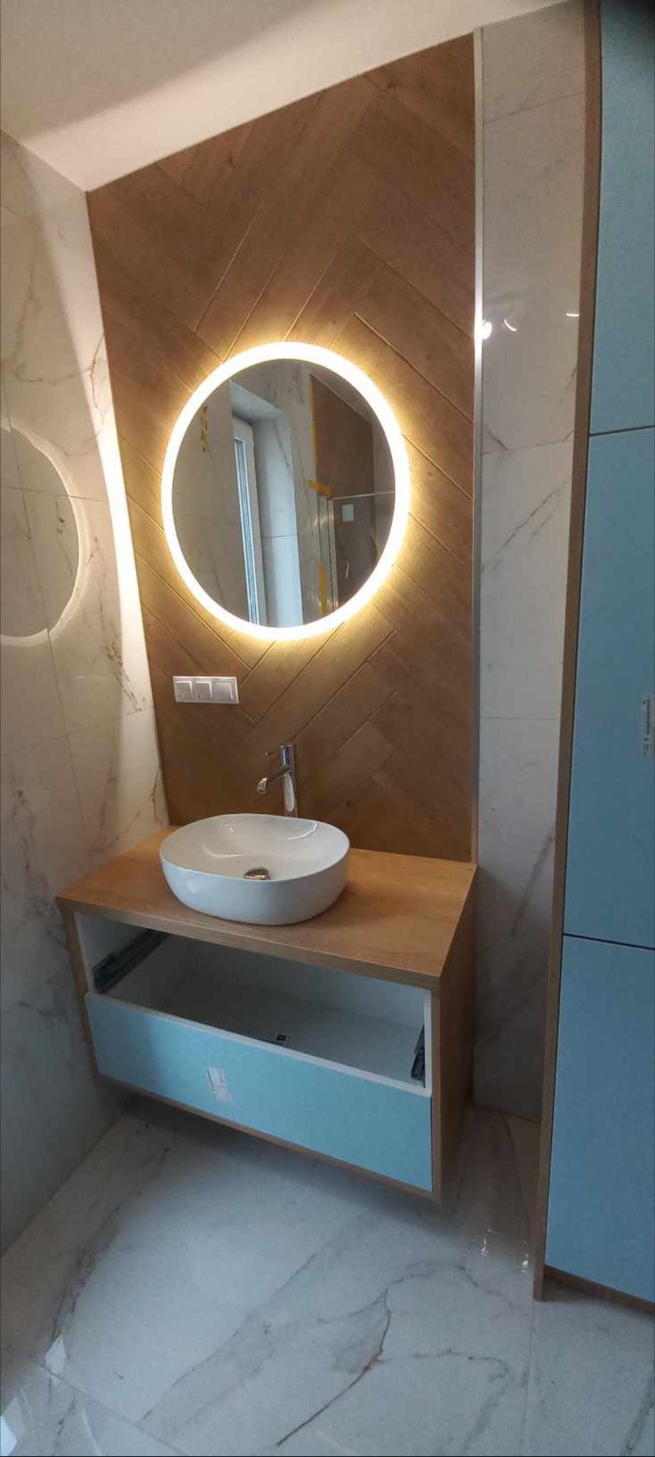 Nowoczesna łazienka z okrągłym lustrem LED, umywalką nablatową i drewnianą ścianą w jodełkę, uzupełniona marmurowymi płytkami podłogowymi.