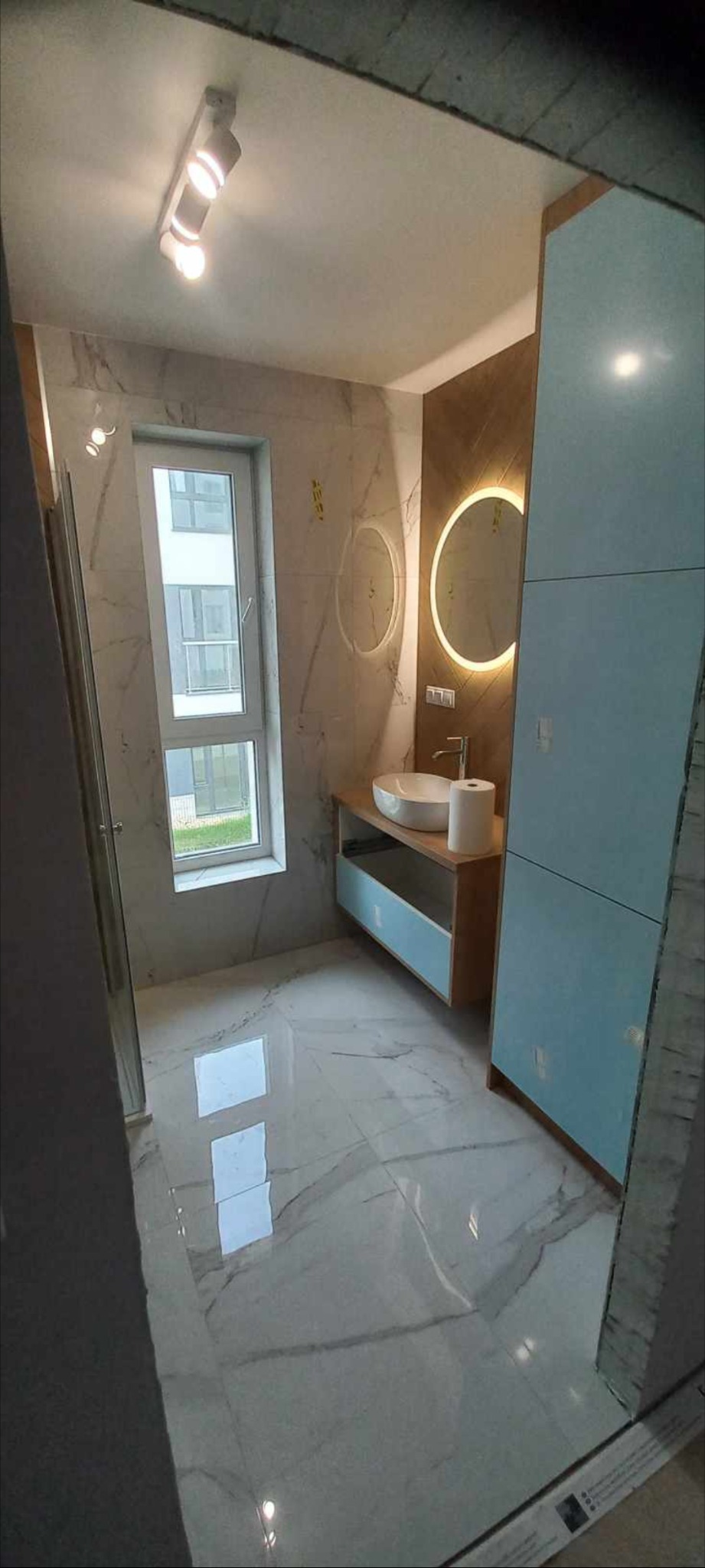 Nowoczesna łazienka z marmurowymi płytkami, okrągłym lustrem z podświetleniem LED, białą umywalką nablatową i szafką z błękitnymi frontami. Widoczne odbicie światła w polerowanych płytkach...