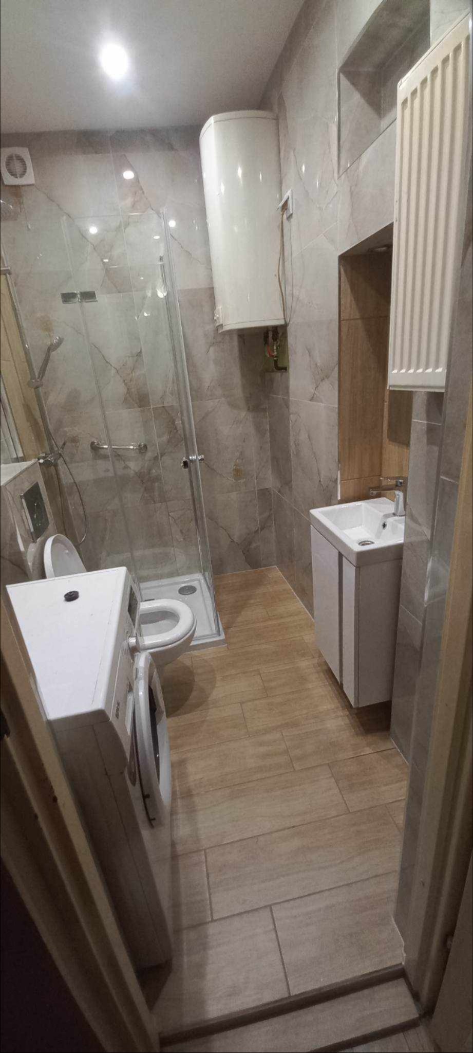 Ukończona łazienka z kabiną prysznicową, umywalką z szafką, toaletą, bojlerem i pralką, wykończona płytkami w odcieniach beżu i szarości, widok zza futryny drzwi.