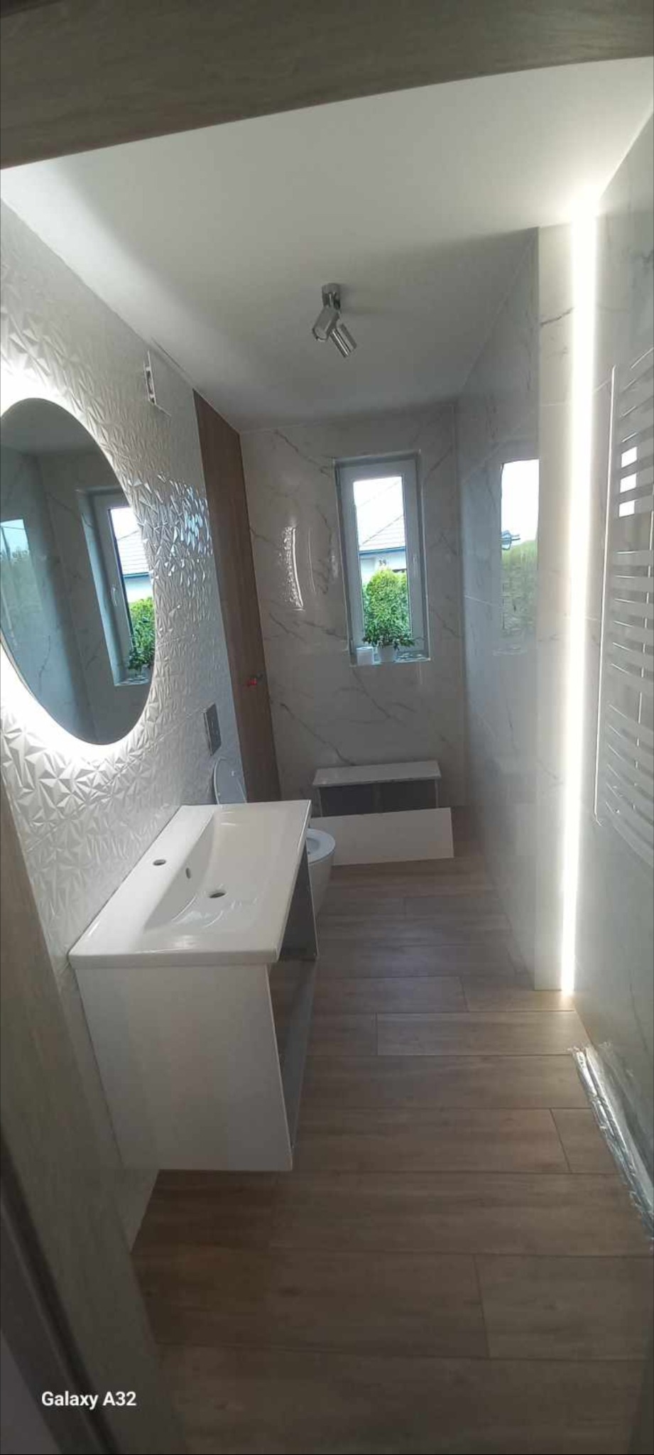 Nowoczesna łazienka z marmurowymi płytkami, drewnopodobną podłogą, okrągłym lustrem z podświetleniem LED i pionowym oświetleniem wzdłuż ściany.