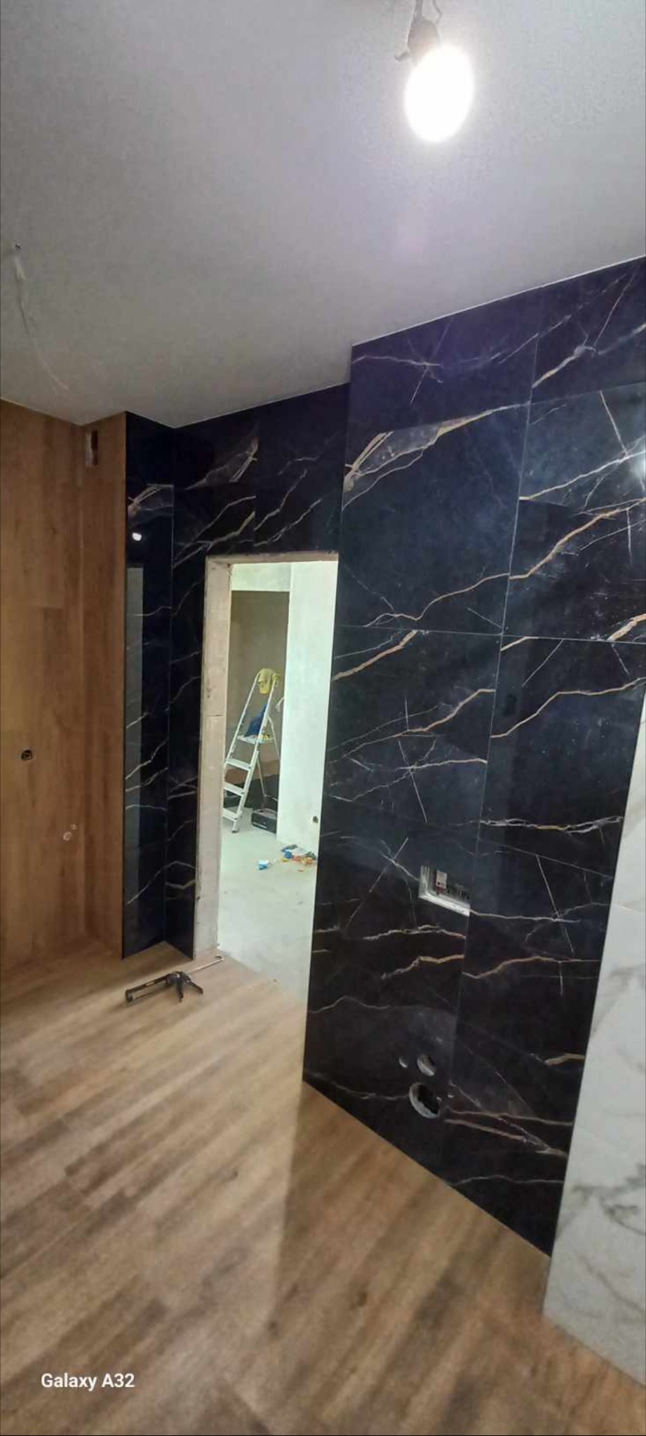 Częściowo wykończona łazienka z czarnymi płytkami imitującymi marmur ze złotymi żyłami na ścianach, drewnopodobną podłogą i widoczną drabiną w przejściu do kolejnego pomieszczenia.