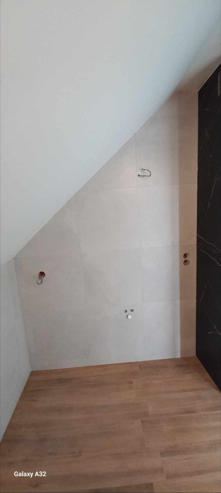 Łazienka w trakcie wykańczania: ściany wyłożone jasnymi płytkami, na podłodze panele imitujące drewno, widoczne wyprowadzenia instalacji wodnej i elektrycznej.