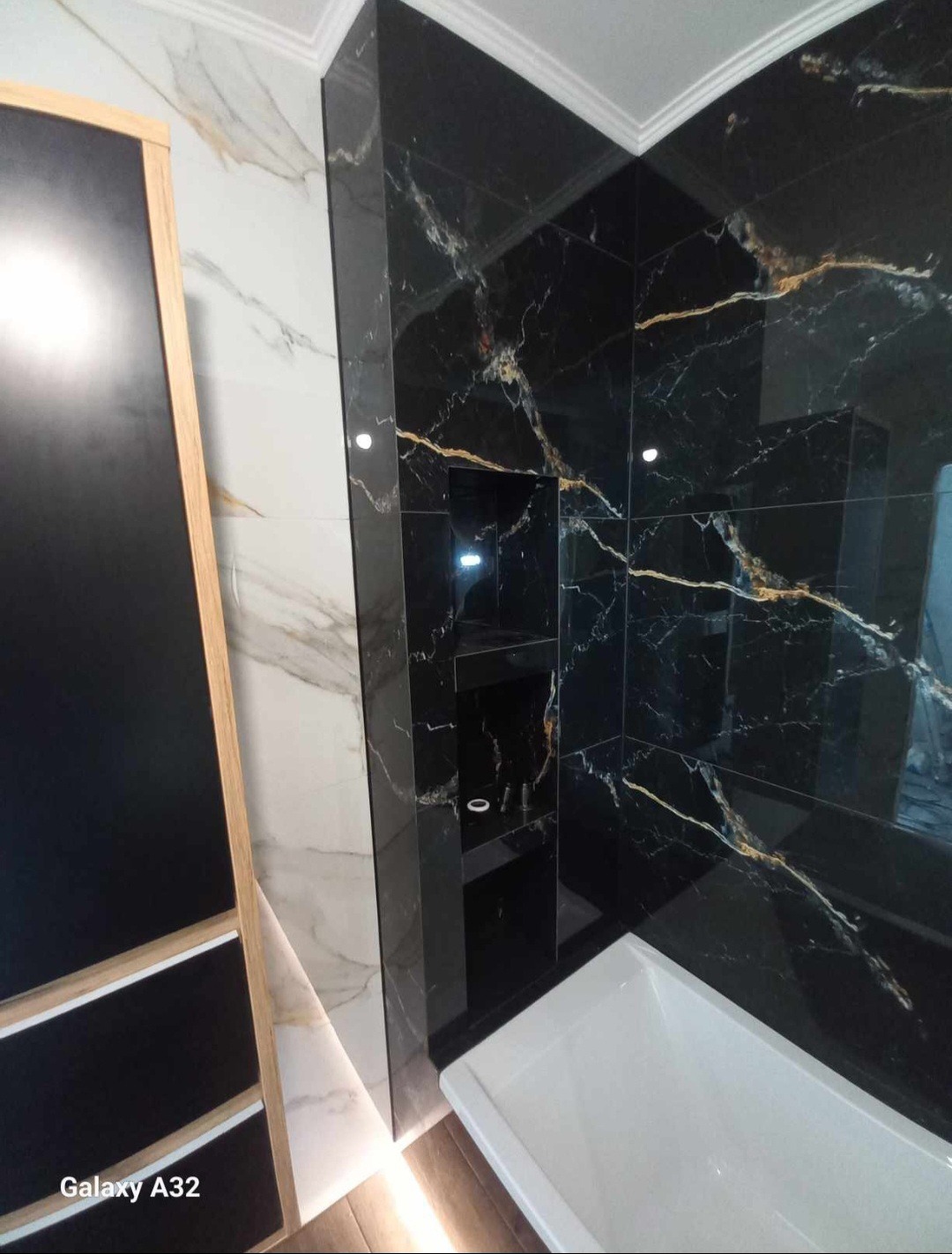 Wykończona łazienka z czarnymi płytkami imitującymi marmur ze złotymi żyłami, wbudowana półka w strefie prysznica, biała wanna, drewnopodobna podłoga, fragment czarnej szafki z drewnianą ramą.