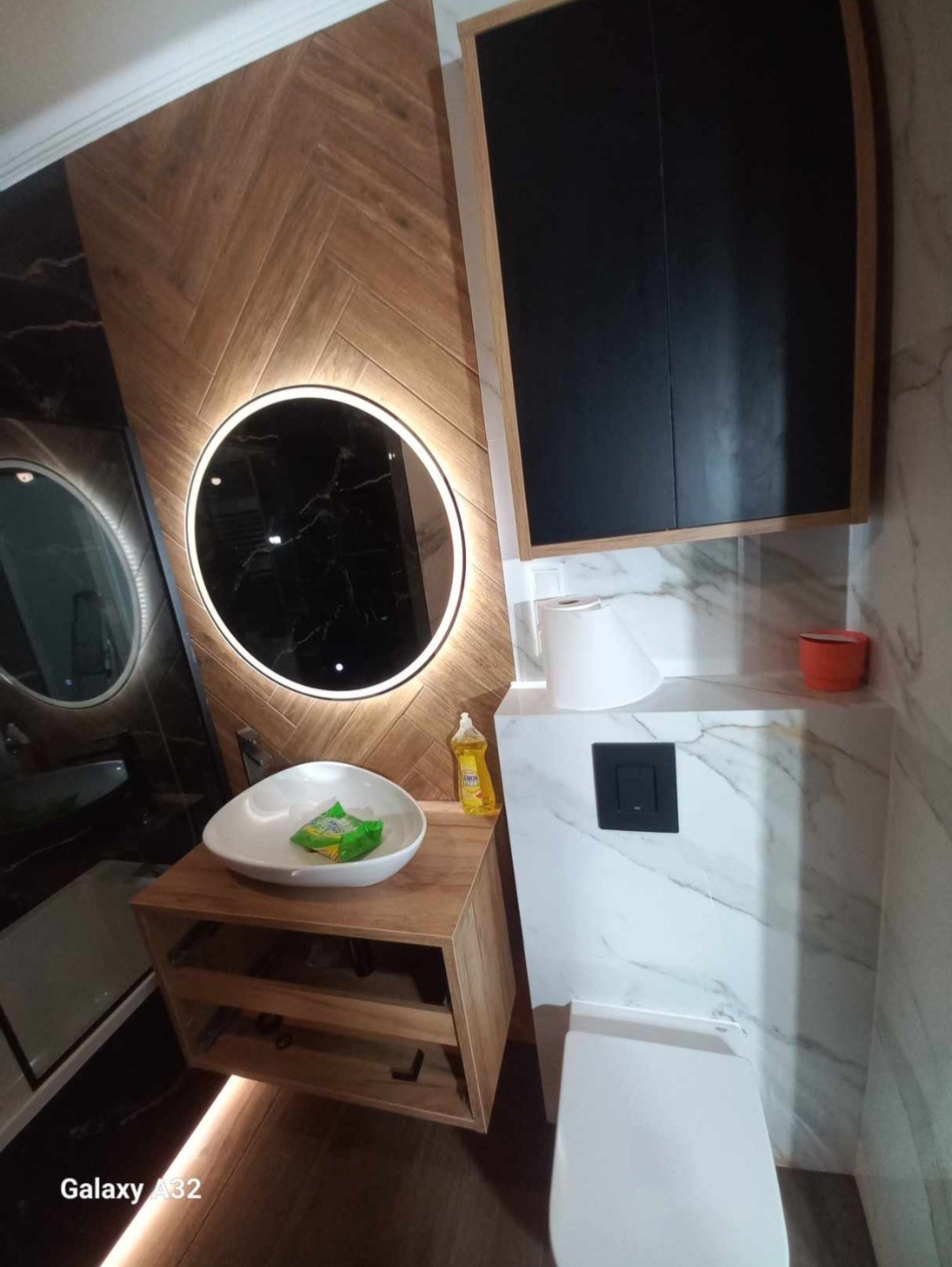 Nowoczesna łazienka z drewnianą ścianą ułożoną w jodełkę, okrągłym lustrem z podświetleniem LED, wiszącą szafką z umywalką oraz toaletą z zabudowaną spłuczką w marmurze.