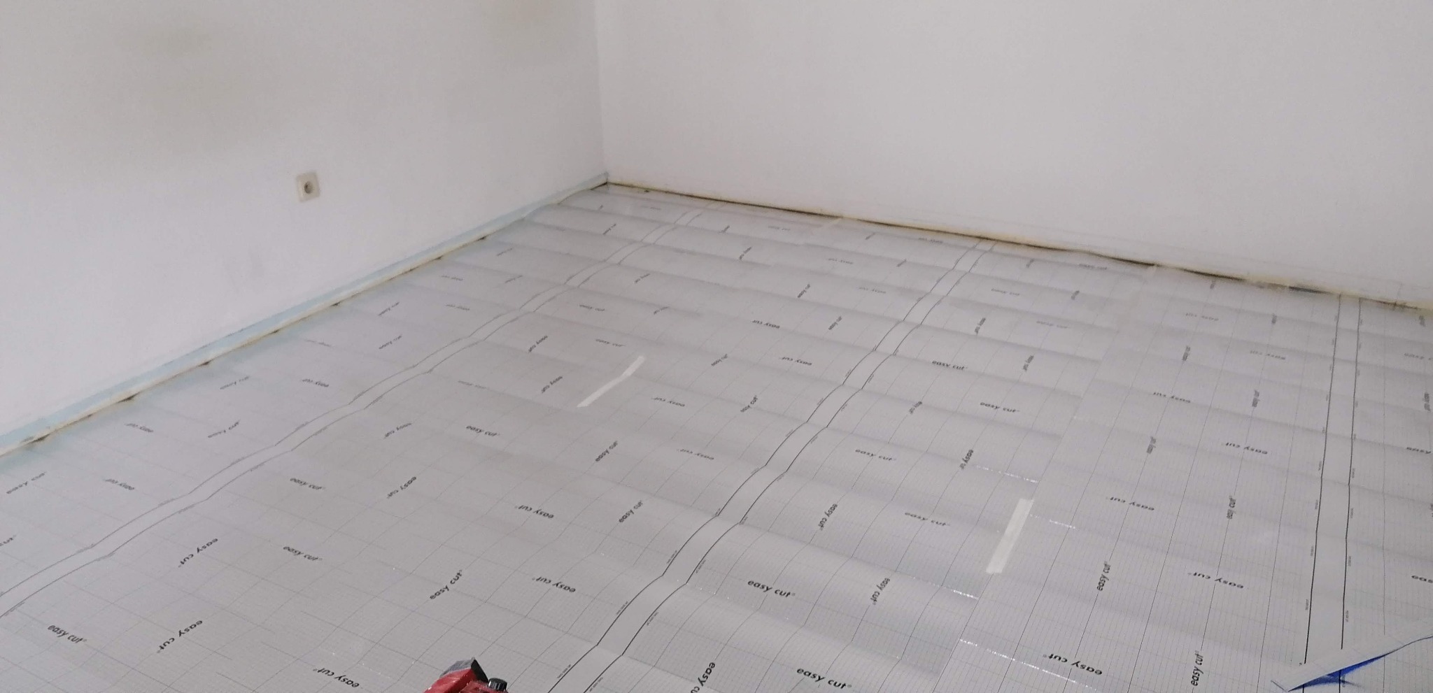 Przygotowanie podłoża pod panele podłogowe: rozłożona folia izolacyjna z nadrukiem siatki i napisem 'easy cut' w rogu pomieszczenia, widoczna listwa przypodłogowa i gniazdko elektryczne.