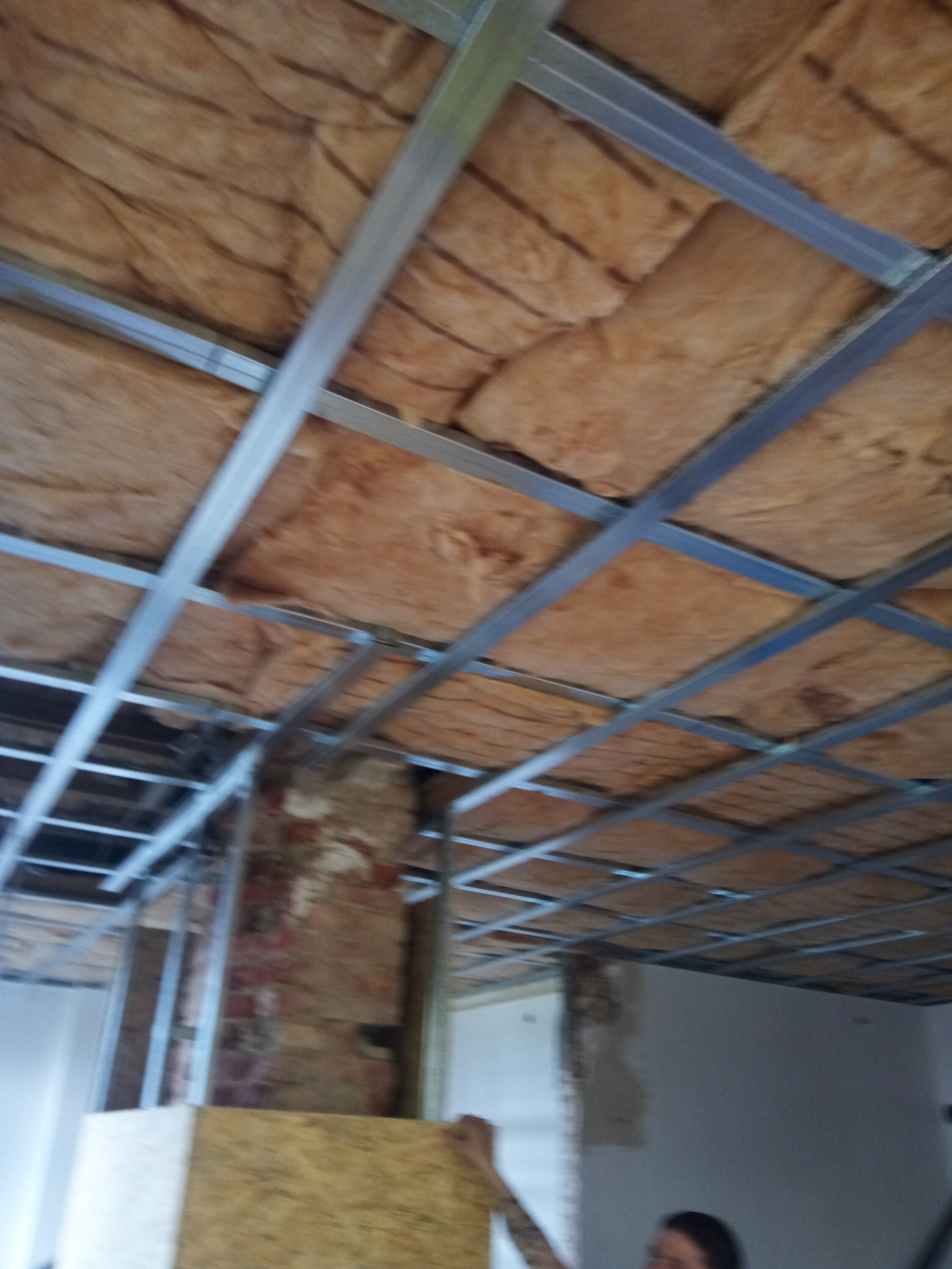 Montaż izolacji z wełny mineralnej na poddaszu, widoczna metalowa konstrukcja nośna i fragment ściany z cegły