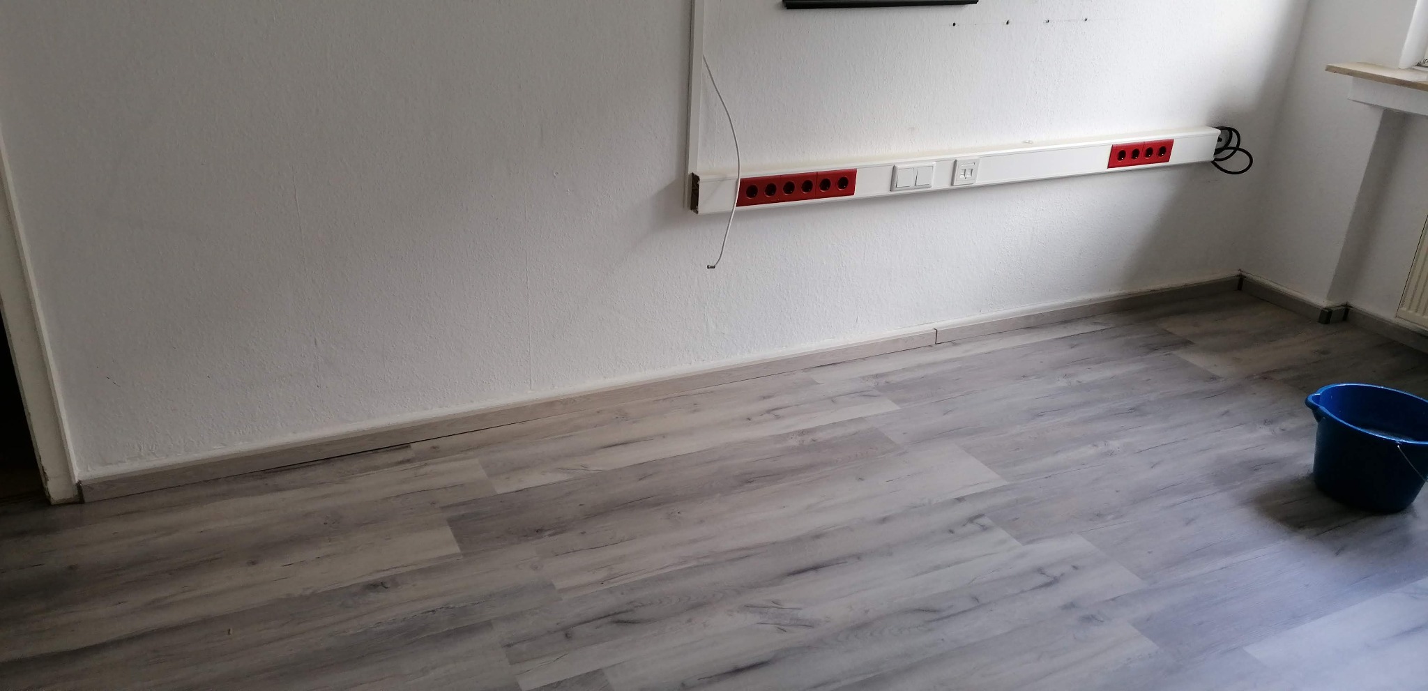 Pusty pokój po remoncie z panelami podłogowymi imitującymi drewno, białe ściany z listwą przypodłogową i listwą z gniazdkami elektrycznymi, niebieskie wiadro w rogu pomieszczenia.