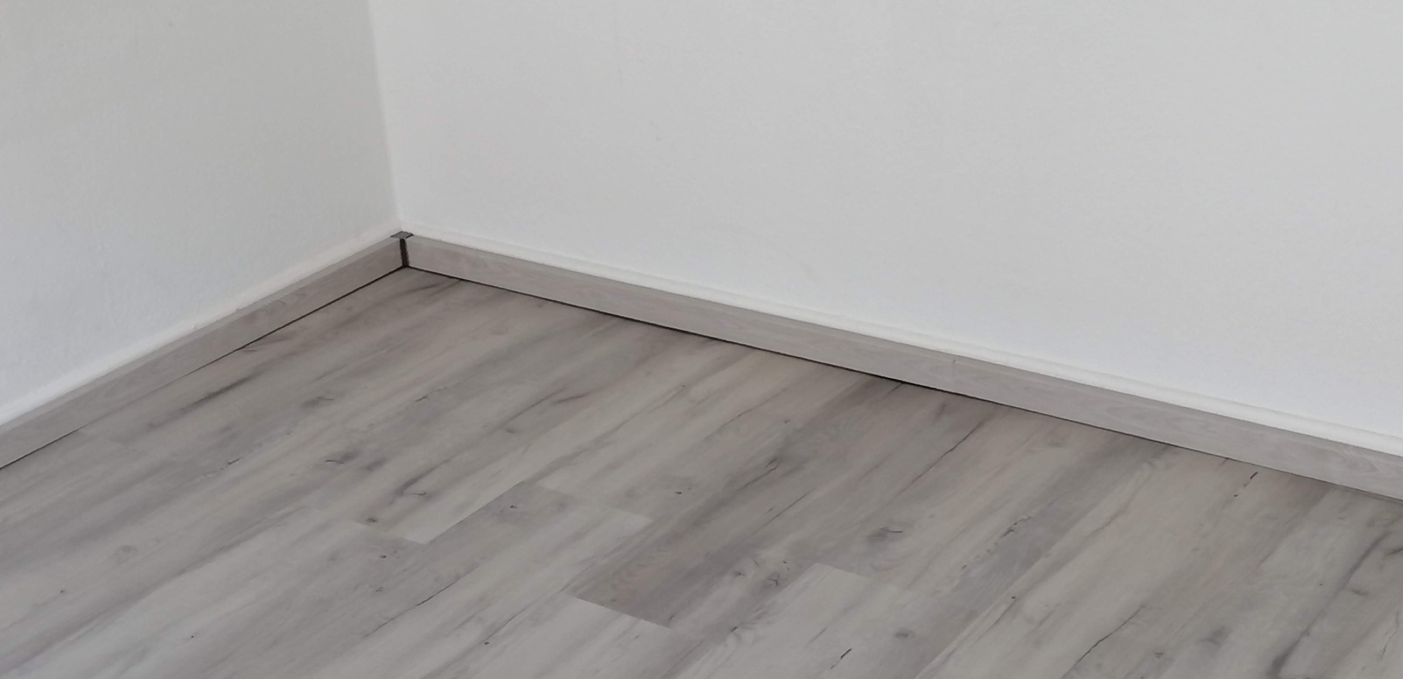 Szare panele podłogowe ułożone w rogu pomieszczenia, widoczne listwy przypodłogowe przy białych ścianach.