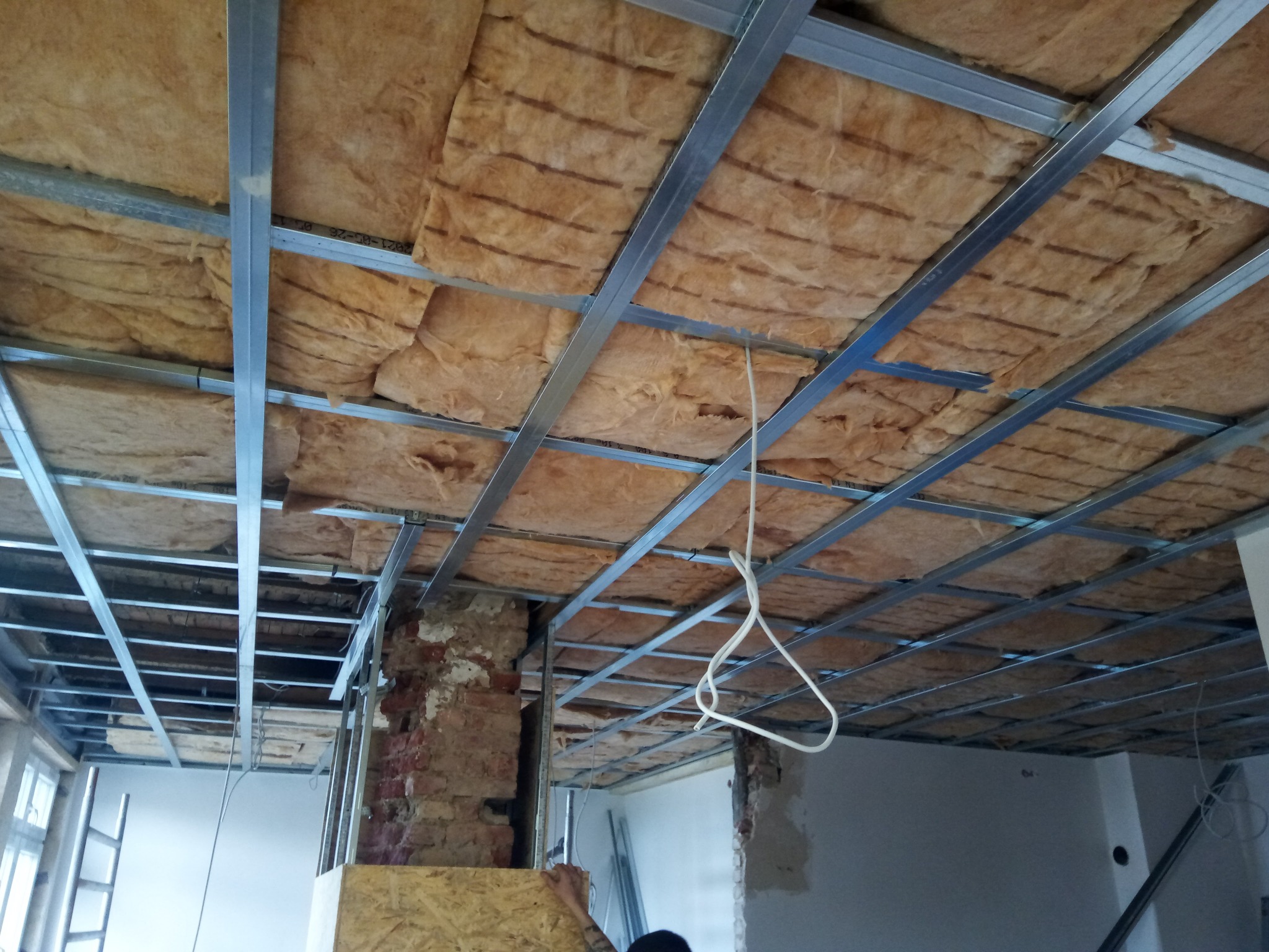Montaż izolacji z wełny mineralnej w metalowym stelażu sufitu podwieszanego, widoczne fragmenty ceglanej ściany i przewody elektryczne.