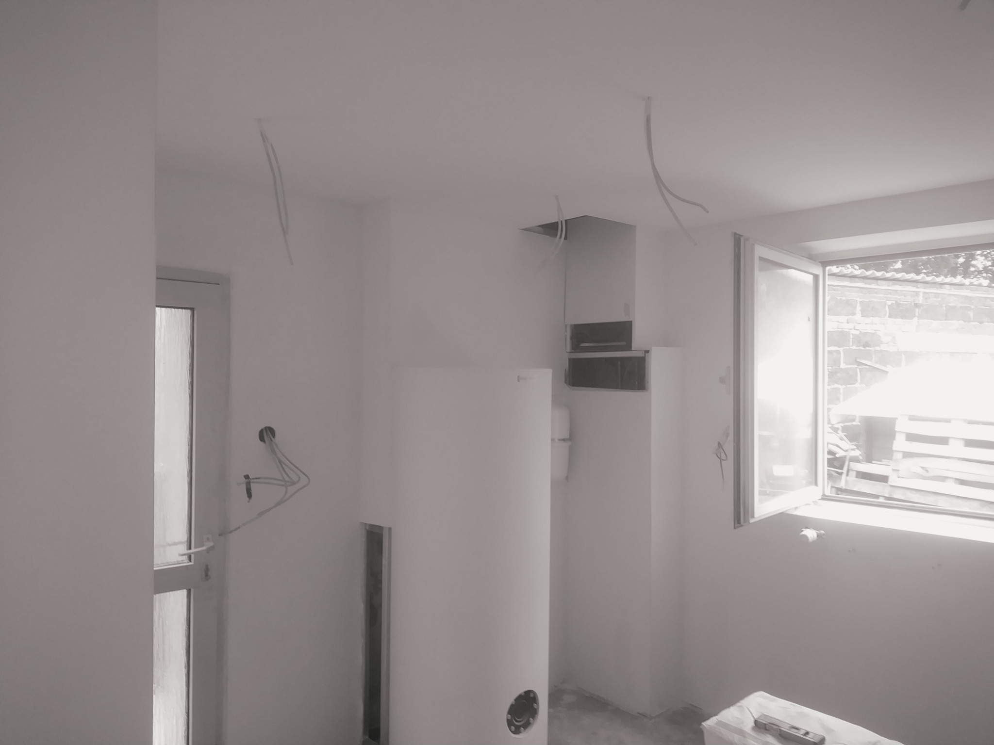 Wnętrze pokoju w trakcie prac: białe ściany, wiszące przewody, bojler, otwarte okno z widokiem na palety na zewnątrz, poziomica na podłodze.